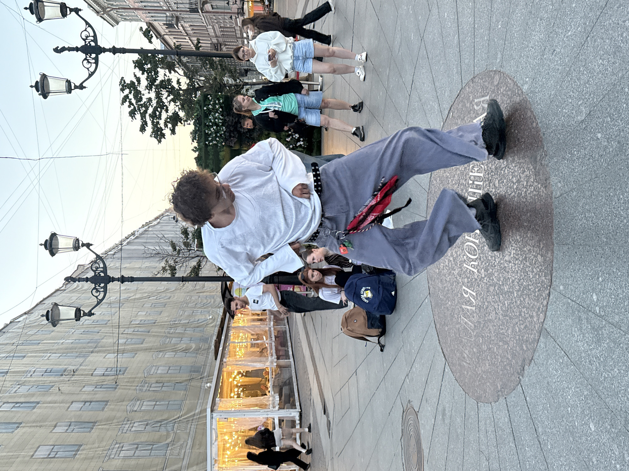 The image captures a group of people on a street, likely in Saint Petersburg, Russia. The main subject is a person in the foreground, dressed in a white long-sleeved shirt and light grey pants, in a dynamic pose as if dancing or performing. They are standing on a circular stone inlay on the pavement, which features visible text.
Behind the main subject, several other individuals are standing, observing. Some are dressed casually in shorts and t-shirts, while others are in hoodies. A young woman in a white hoodie and denim shorts stands out, holding a phone. Another person in a turquoise shirt and denim shorts is also visible. Bags, including a navy blue backpack, are placed on the ground near some of the bystanders.
The scene appears to be outdoors on a paved street with a building facade in the background, partially covered by a large banner that mimics windows. Streetlights with multiple lamps are mounted on ornate poles. In the distance, a row of stalls or a covered walkway with illuminated lights can be seen, suggesting a lively area, possibly a market or an outdoor dining space.
The lighting suggests it is late afternoon or early evening, with a soft, diffused light. The sky is visible in the upper portion of the image, appearing overcast or hazy. The overall mood seems relaxed and casual, with people gathered in a public space.
The text visible on the circular inlay on the ground appears to be in Cyrillic script. While not entirely clear, portions of it seem to read "МАЯ КО..." and possibly other characters. The overall setting evokes a sense of urban life and a public gathering.