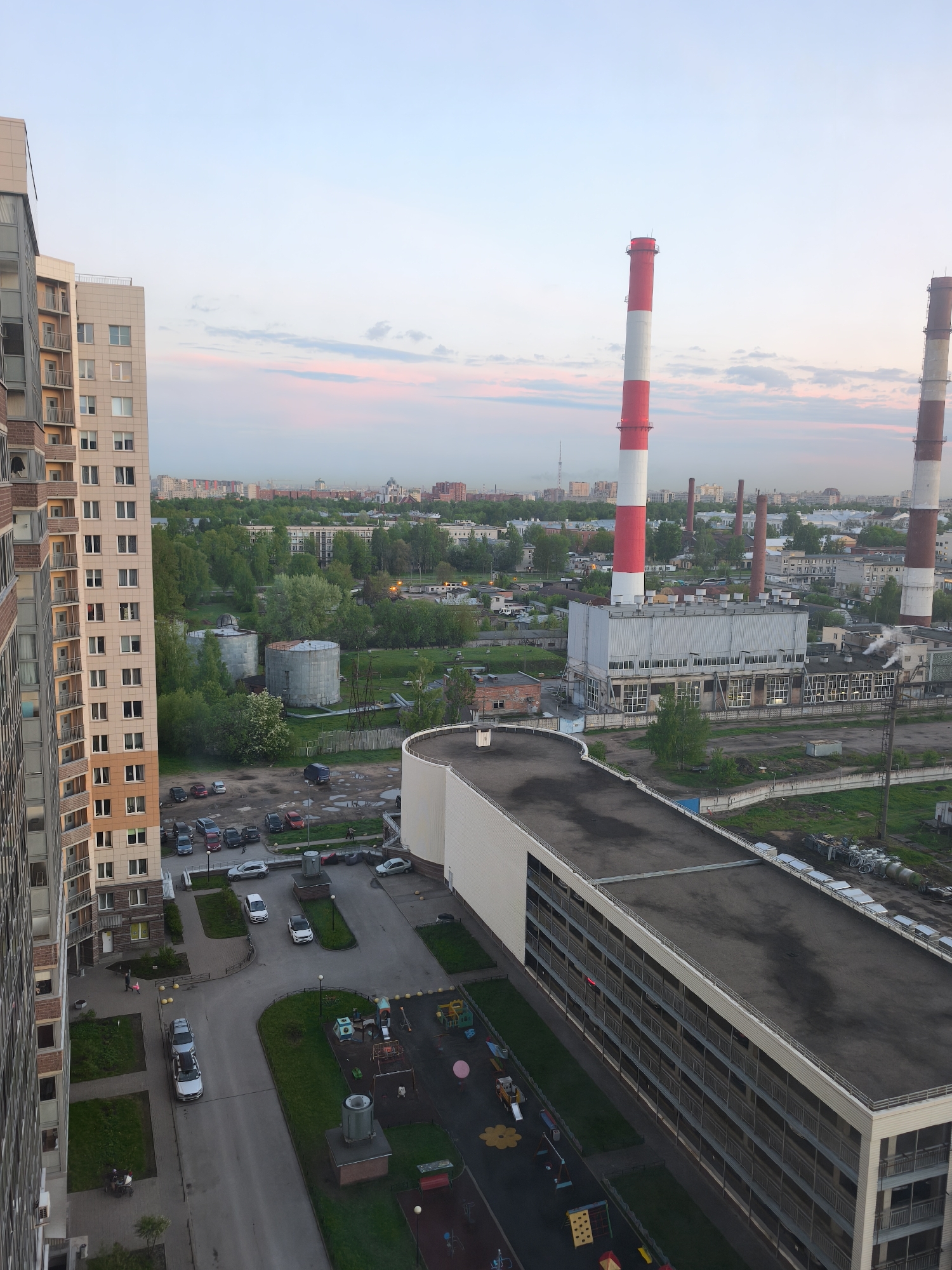 The image depicts a high-angle view of an urban scene, likely taken from a high-rise building in Saint Petersburg, Russia. A tall apartment building with light-colored balconies is visible on the left. In the foreground, there's a parking lot with several cars and a playground with various equipment. A multi-story building with a flat roof extends horizontally across the scene. In the distance, there are industrial chimneys, some painted with red and white stripes, rising above a mix of buildings and green spaces. The sky is pale with pink hues, suggesting either sunrise or sunset. Overall, the scene presents a mix of residential, industrial, and recreational elements within a densely populated urban environment.