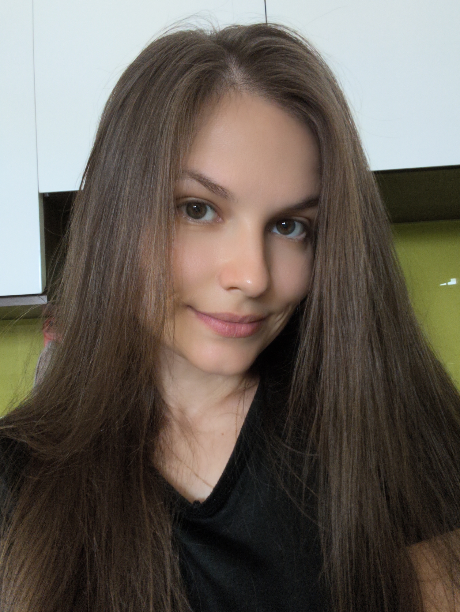 A young woman with long, brown hair is looking directly at the camera. She is wearing a black shirt. The background is blurry, with hints of white cabinets and a lime green wall. The scene appears to be indoors, possibly a kitchen or living area. No specific location in Bac Nha Trang Ward, Vietnam, can be determined from the image.