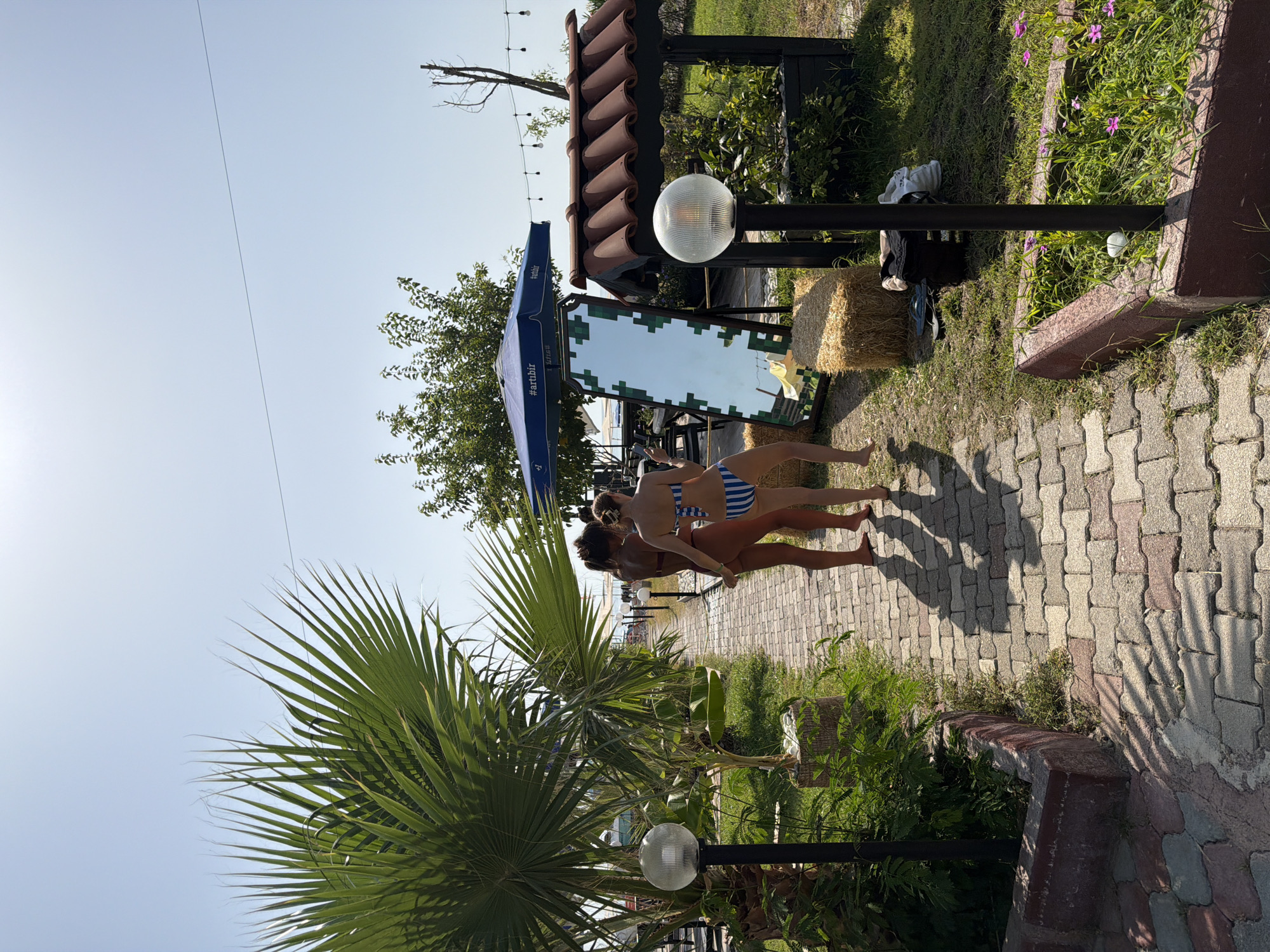 In Kemer, Turkey, a paved walkway leads through a grassy area with a palm tree on the left. Two women in bikinis stand on the walkway, facing away from the camera, with one appearing to be adjusting the other's bikini. To the right, a large mirror with a pixelated frame stands propped up, reflecting the sky and a blue umbrella with "artibir" text. Behind the mirror are bales of hay and a structure with a tiled roof. In the foreground, planters hold greenery and small pink flowers. Outdoor lighting fixtures are visible along the walkway and in the background.