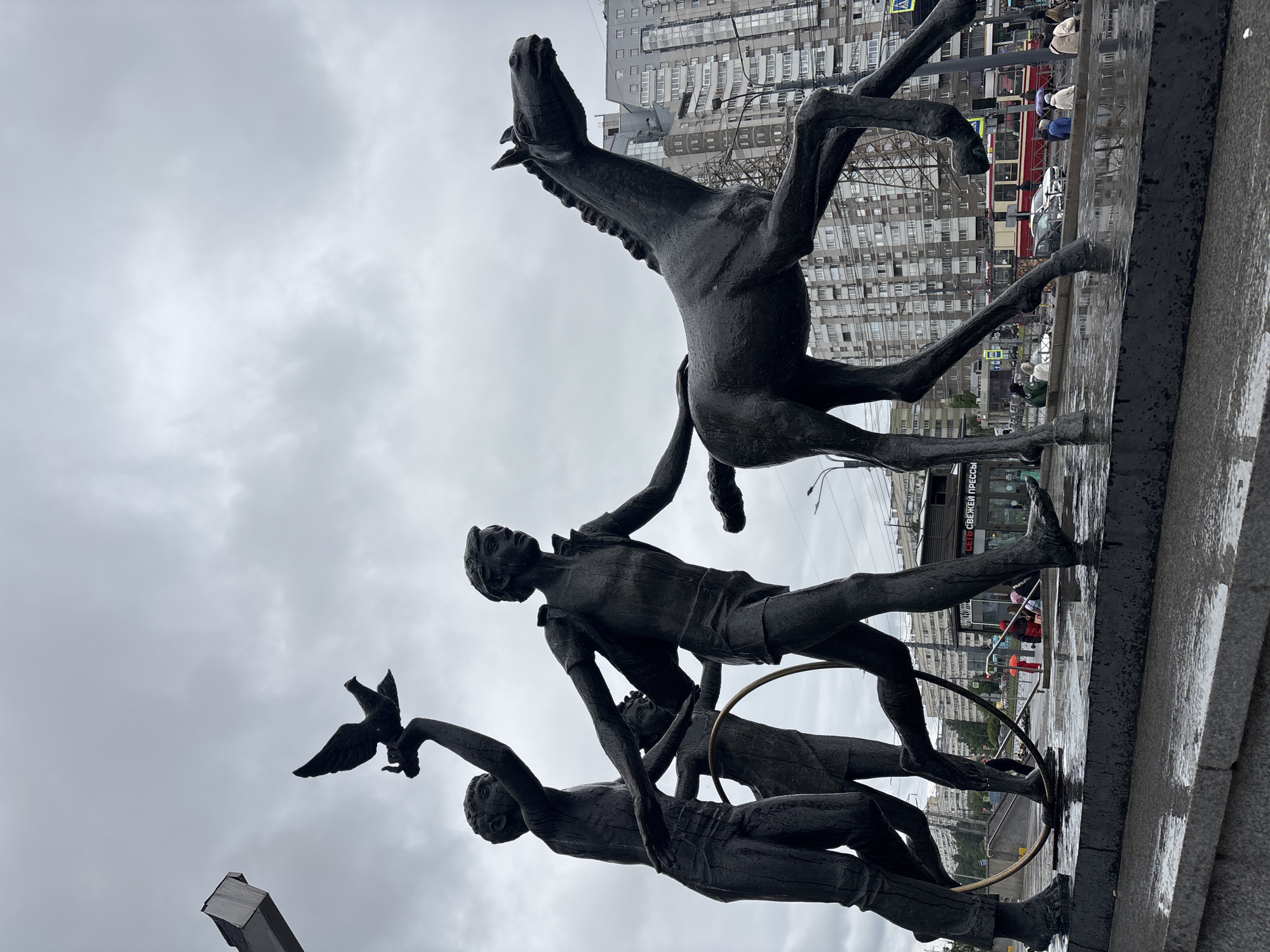 The image shows a statue of a horse and three men with a bird. The statue is located in the city of Saint Petersburg, Russia. The horse is rearing up, and the men are positioned underneath. One man is holding a bird. One man is holding up the horse's tail. A ring or hula hoop is positioned behind the central figure. The background shows a tall apartment building on an overcast day. The ground is wet.
