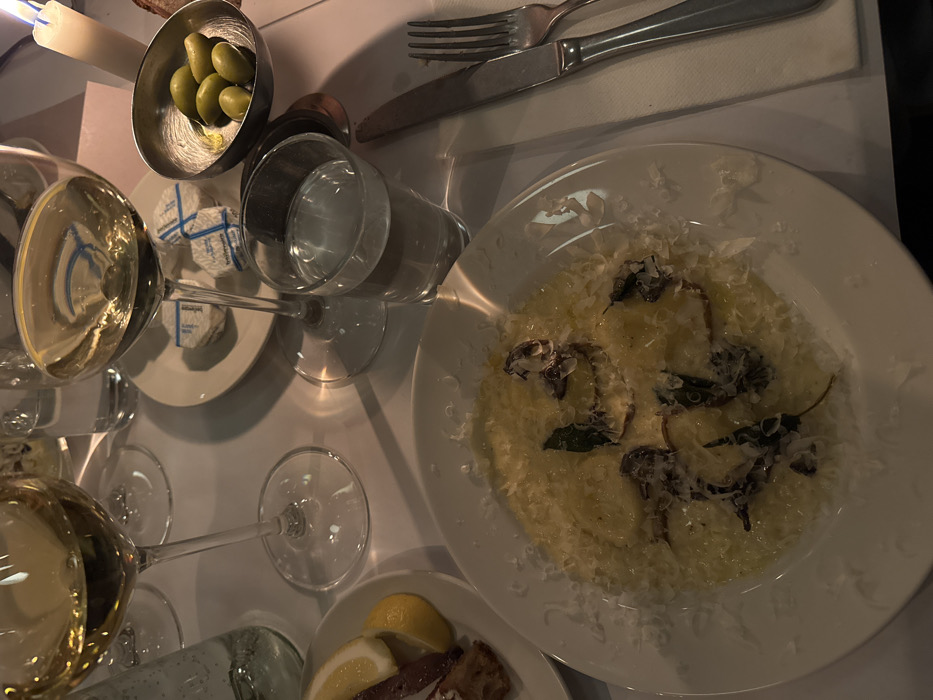 A tabletop with various dishes includes a white plate of pasta adorned with shredded cheese and herbs. Alongside it, a smaller plate presents lemon wedges and dark slivers. A metal bowl holds green olives, while another plate displays wrapped cheese portions. A knife and fork rest on a napkin. Glassware includes a filled wine glass, a water glass, and empty wine glasses. A candle adds subtle lighting to the scene.