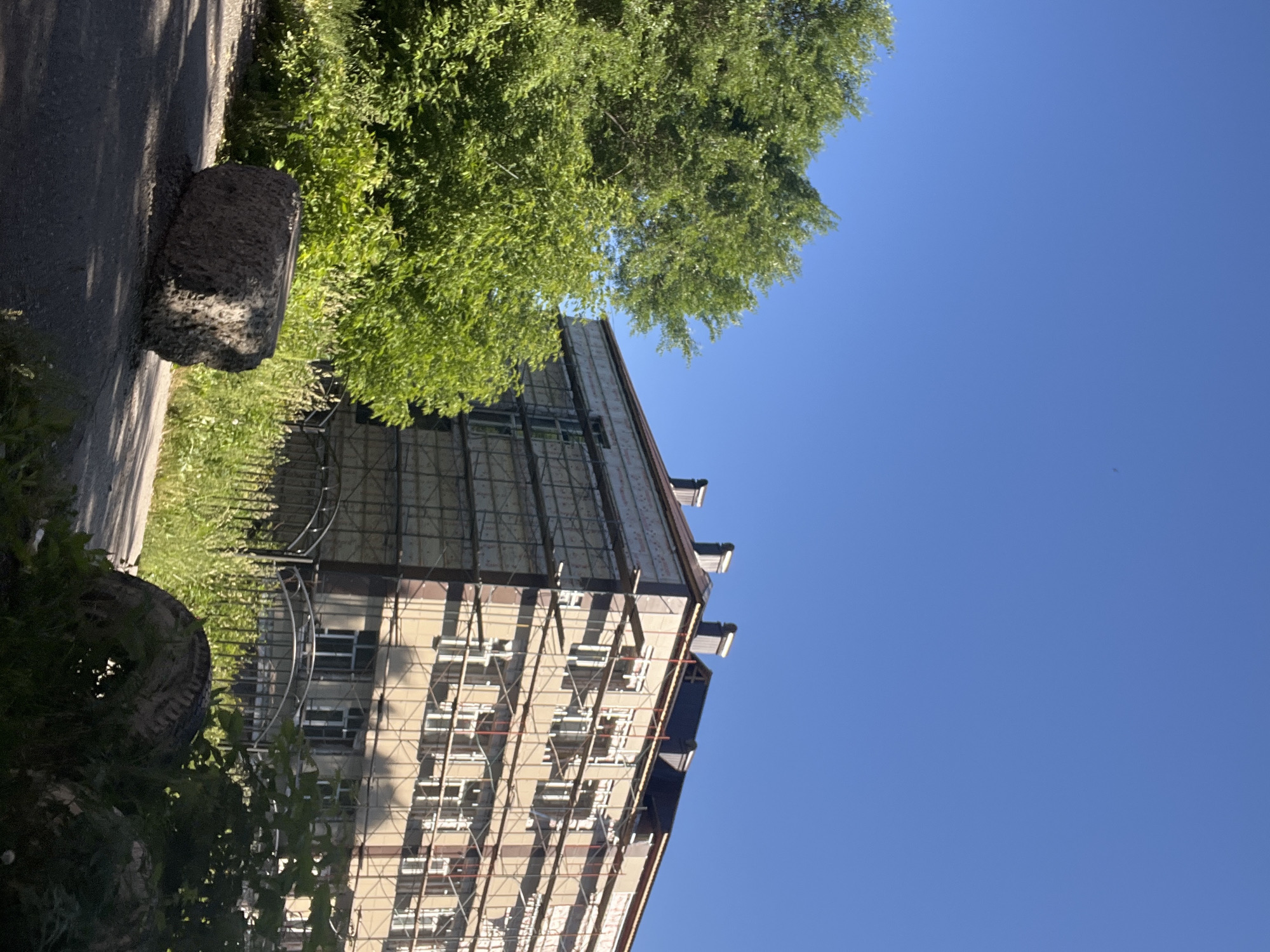 The image depicts an outdoor scene in Kiselyovsk, Russia, under a clear blue sky. In the foreground, a grey, rectangular concrete block rests on an asphalt path. To the right of the block, a worn tire is partially obscured by green foliage. Behind these elements, a building undergoing renovation stands, encased in scaffolding. The building features multiple floors with windows, and its facade is a light brown color. To the left of the building, a lush green tree with dense foliage extends upwards. The overall environment suggests a daytime setting with bright sunlight.