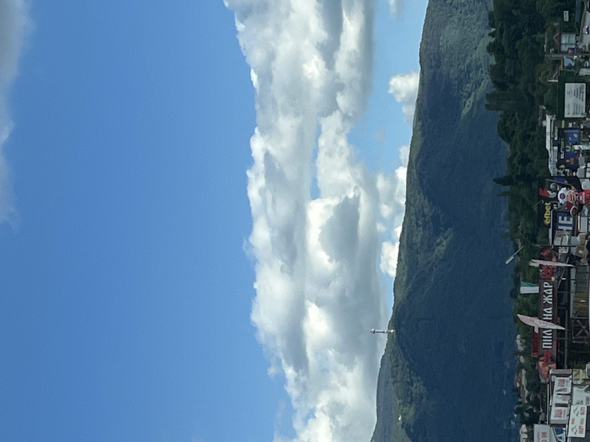 In the city of Unknown, Unknown, a wide-angle, vertical shot captures a mountainous landscape under a bright blue sky dotted with fluffy white clouds. A tall, slender tower with a red and white structure at its apex stands on the peak of a densely forested mountain. Below the mountain, a strip of urban settlement is visible, featuring a collection of buildings and billboards. One prominent billboard displays text in Cyrillic script that reads "ПИП НА ЖАР". Another billboard shows the "efbet" logo. A few vehicles are present on a road in the foreground. The scene is bathed in daylight, suggesting it was taken during the day.