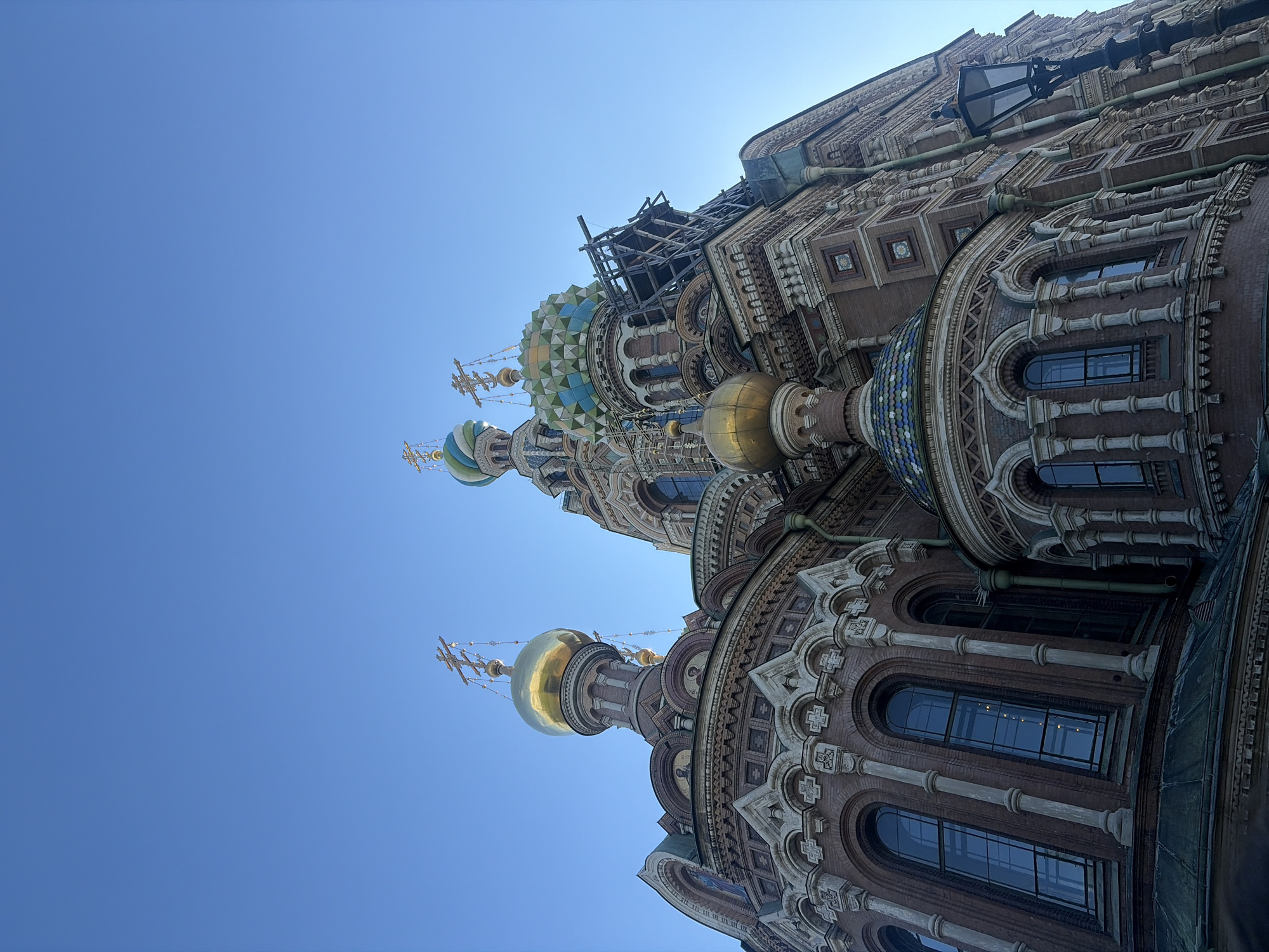 This is an upward-facing, wide-angle shot of the exterior of the Church of the Savior on Spilled Blood in Saint Petersburg, Russia. The clear blue sky fills the background, suggesting a bright, sunny day. The architecture of the church is intricate and colorful, featuring onion domes, mosaic patterns, and decorative brickwork. Several domes are visible, with the largest being a golden onion dome topped with a cross. Another dome nearby is partially covered in blue and green mosaics, and another is a patterned green and white. The building's facade displays numerous arched windows and detailed ornamentation with a light-colored stone against a darker brick. Some scaffolding is visible on the upper section of the church, indicating ongoing construction or restoration work. A dark lamppost is mounted on the side of the building. The low angle and tilted perspective emphasize the grandeur and height of the church. No people are visible in the frame.
