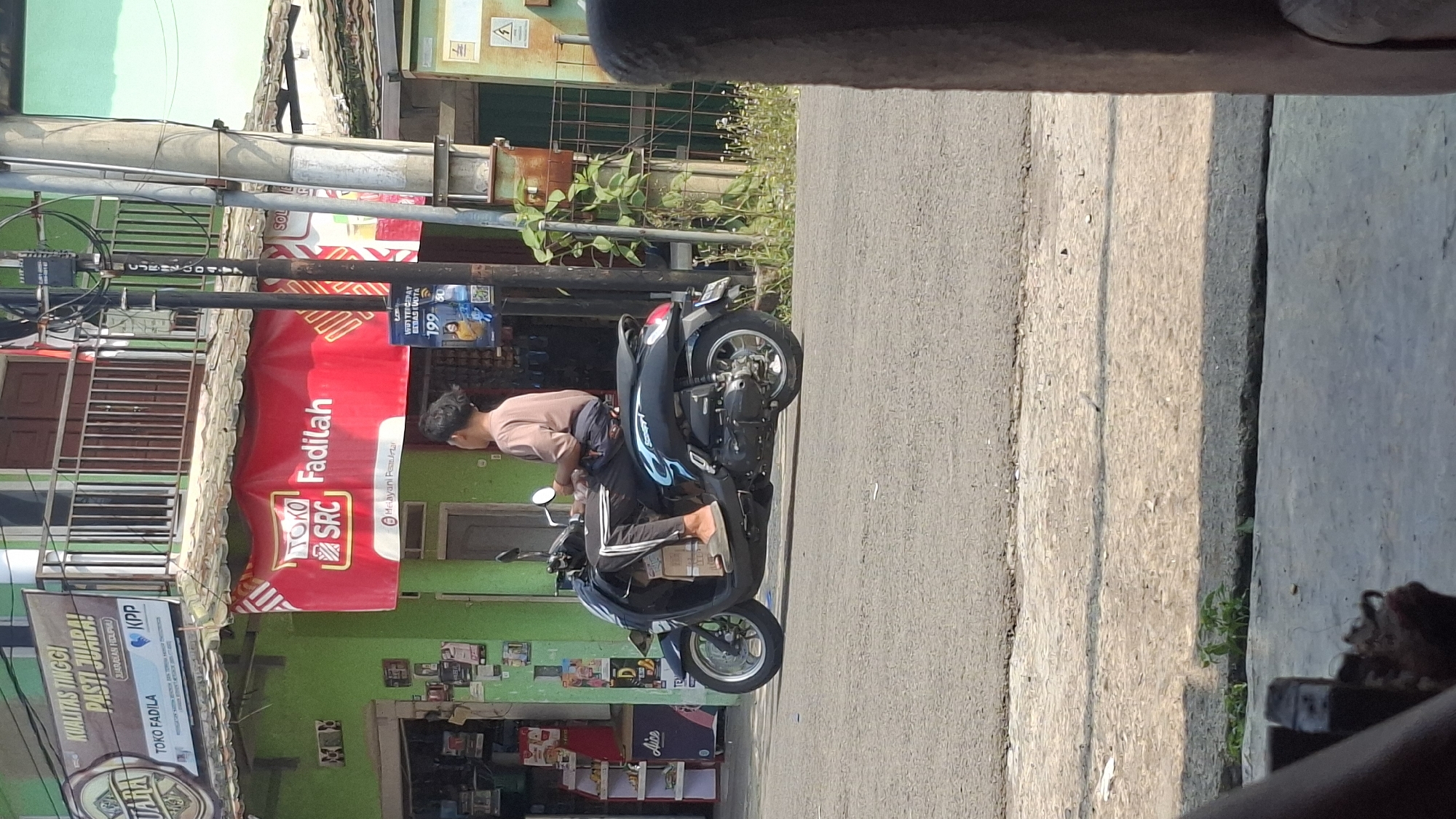 This is a medium shot, eye-level, outdoor, daytime photograph taken in Cilaku, Indonesia. A young person wearing a light brown t-shirt and dark pants with white stripes is riding a black scooter on an asphalt road. The scooter has blue and white graphics on the side and a license plate. The person is looking to the right, with their right foot resting on the road.
In the background, a bright green building with a tiled roof is visible. A red banner with white text, including the words "Toko Fadilah" and "SRC", is displayed on the building. There is also a metal railing and a utility pole with wires. Further to the right, there is a partially visible wooden structure and greenery. The foreground shows a concrete sidewalk and road.