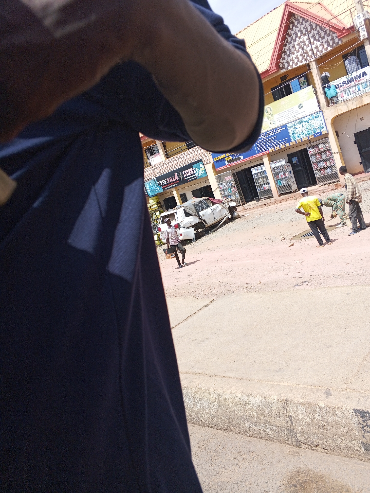 The image captures an outdoor street scene in Jos, Nigeria, focusing on the aftermath of a car accident. In the immediate foreground, a person's elbow and dark blue clothing are visible, indicating the photo was taken from a low angle, possibly by someone walking or standing close to the observer.
The central focus of the midground is a severely damaged white sedan, its front end crumpled, resting at an angle on the unpaved, dirt and gravel road. Several men are gathered nearby, observing the scene. One man in a bright yellow t-shirt and dark pants stands with his hands on his hips, looking towards the damaged vehicle. Another man in a plaid shirt stands further back, also facing the car. A third individual, possibly in work attire, is seen bending over near the car. A man carrying a black object walks away from the car towards the foreground.
Lining the street are multi-story commercial buildings, painted in shades of yellow and brown, with distinctive red-tiled roofs. Storefronts display various goods, and several signs are visible. Text detected via OCR includes: "EKESON," "TYREVILLA," "TYRES & WHEELS," and "www.tyrevilla.com" above a shop with stacked tires. Another sign reads "Mobil," and further down, "DISMWA Fashion Home" and "Ready to Wear" are visible on a building that also mentions "AMERICAN AUTO PARTS" and "WE DEAL ON MOTOR SPARE PARTS."
The scene is brightly lit by natural daylight, suggesting it is mid-morning or early afternoon, with clear skies. Shadows are visible on the ground and buildings. The general atmosphere appears calm, with onlookers observing the damaged vehicle rather than engaging in frantic activity, implying the immediate emergency has passed.
