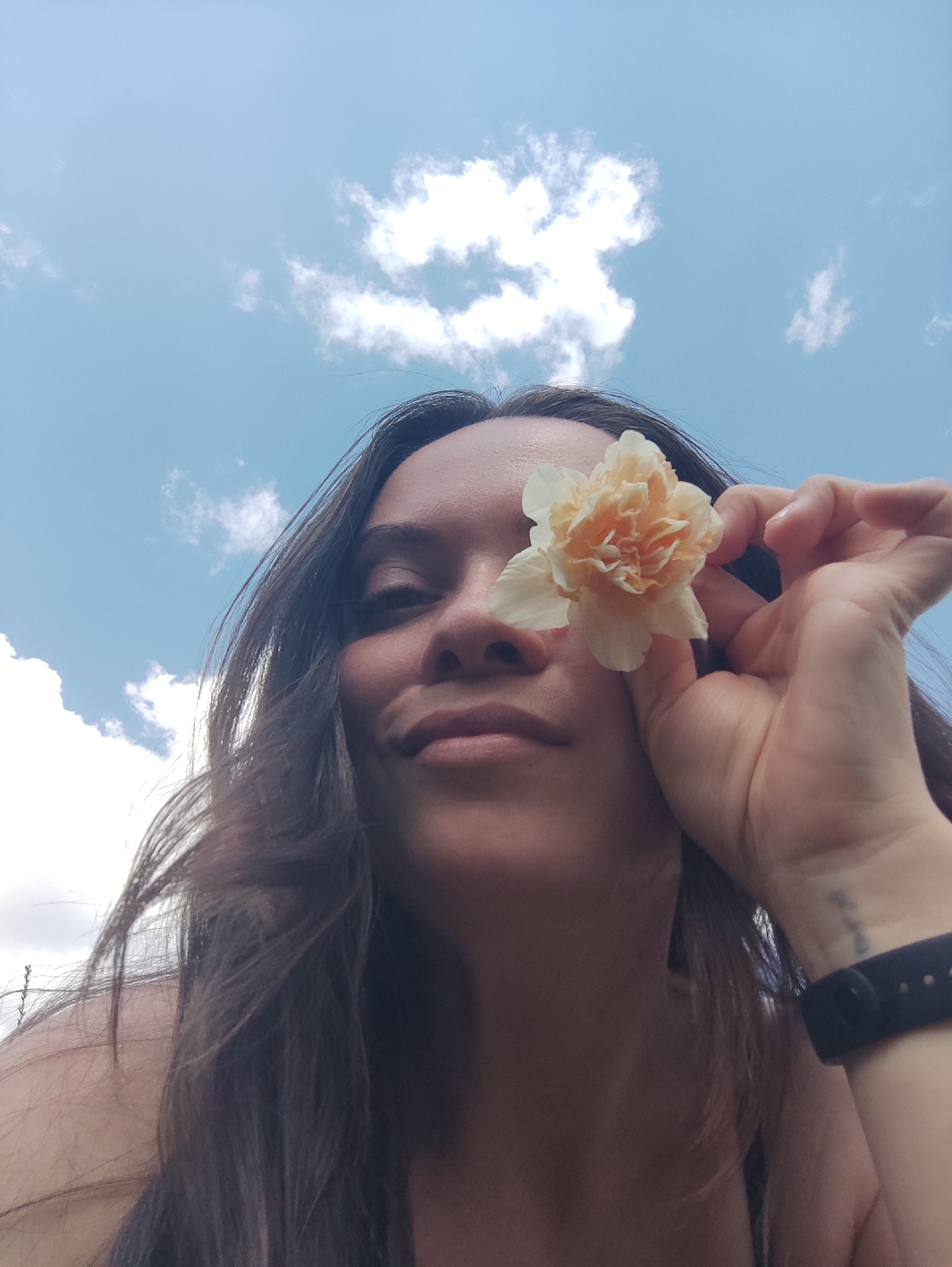 A woman is holding a pale orange flower in front of her eye. The photo appears to be taken from a low angle, looking up at the woman and the sky, which is blue with white clouds. She has dark hair, and her skin tone is light. She is wearing a black fitness tracker on her wrist. It is not possible to determine the location as Фоки, Russia from the provided image.