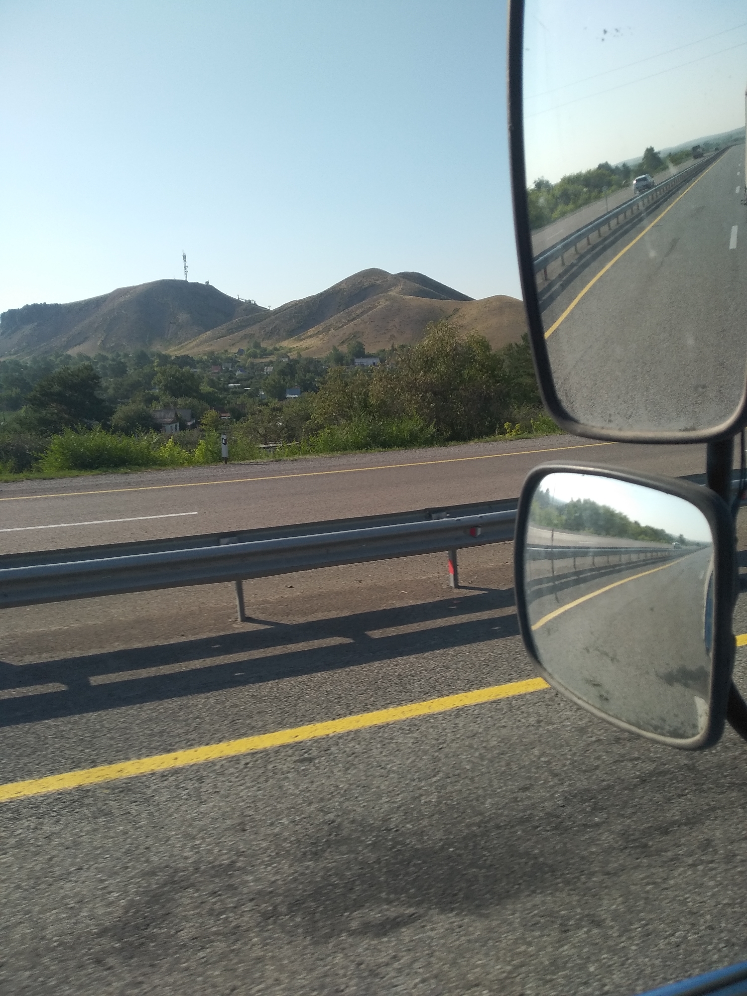 The image shows a view from a vehicle traveling on a highway in Termirtau, Kazakhstan. In the foreground, the asphalt road is marked with a yellow line and a white line. A metal guardrail runs along the side of the road. The right side of the image features two side-view mirrors of the vehicle. The top mirror reflects the sky and a distant, hazy landscape. The bottom mirror reflects the road ahead, showing a few cars and trees lining the highway. In the background, rolling hills are visible, with a prominent radio tower on one of the hilltops. The sky is clear and blue. The scene appears to be set in a rural or semi-rural area on the outskirts of Termirtau.