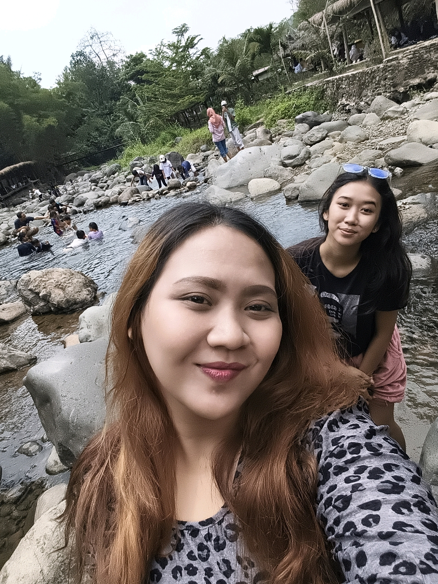 In Babakan Madang, Indonesia, a group of people are enjoying leisure time at a rocky river. Two young women are in the foreground, posing for a selfie. The woman on the left is closer to the camera, her upper body and face visible. She has long, auburn hair with blonde highlights and is wearing a grey leopard-print top. She is smiling and looking towards the camera. To her right, slightly behind her, is another young woman with long, dark hair. She is looking at the camera and wearing a black t-shirt and pink shorts. She has blue sunglasses perched on her head.
In the background, several other individuals, including children and adults, are interacting with the river. Some are wading in the water, while others are sitting on the rocks. The environment is characterized by numerous large, grey rocks scattered throughout the shallow river. Lush green vegetation, including trees and foliage, frames the scene, with a rustic structure, possibly a shelter or cafe, visible in the upper left background. The sky is overcast.