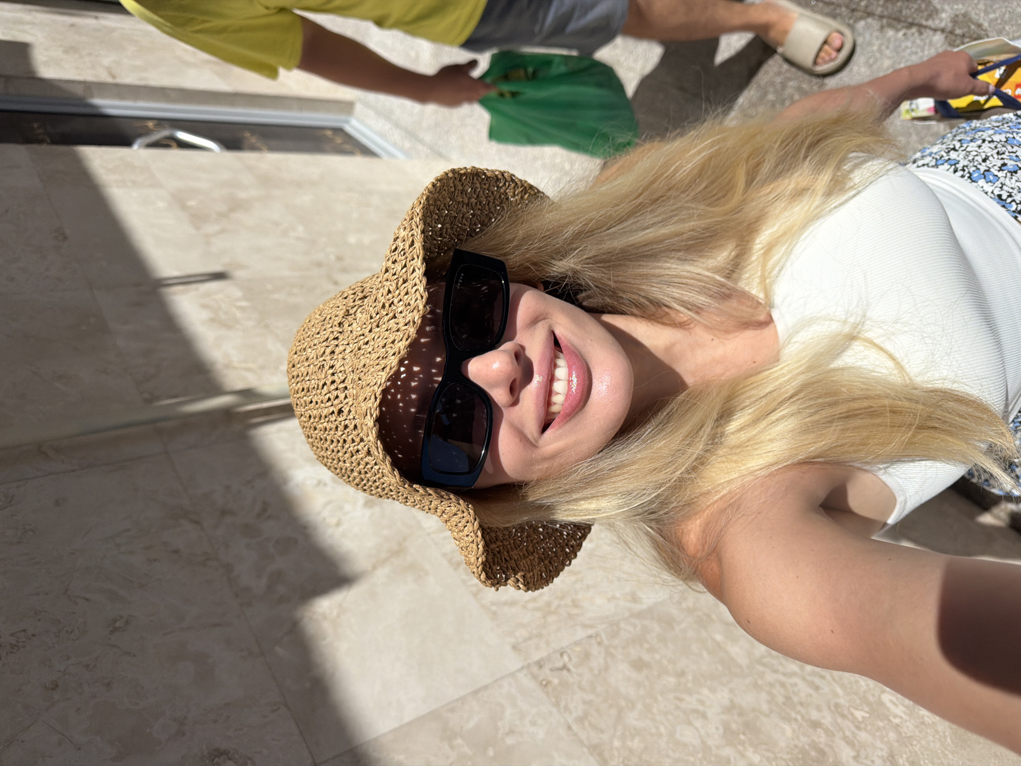 In Budva, Montenegro, a woman with blonde hair, wearing a straw hat and sunglasses, smiles at the camera. She is wearing a white tank top and a patterned skirt. In the background, a person in a yellow shirt and shorts walks away, carrying a green shopping bag. The scene appears to be outdoors, with a tiled wall and shadows from the sun.