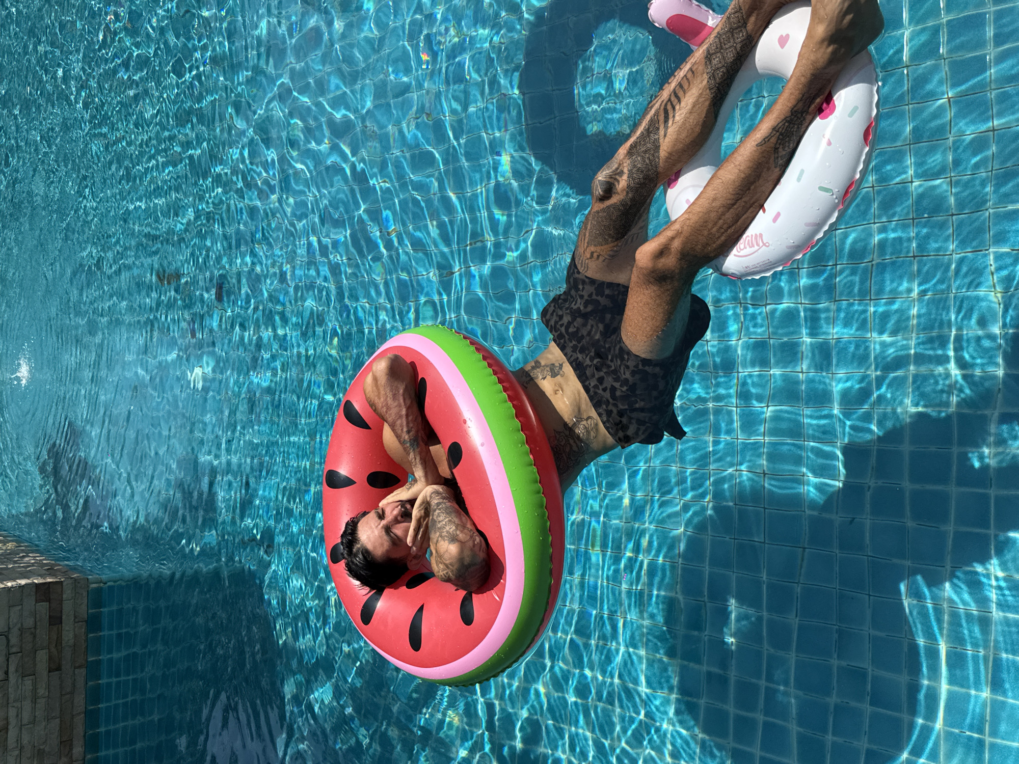 A man is floating in a swimming pool in Pattaya City, Thailand. The pool is tiled with small, square, light-blue tiles. The man is floating on two inflatable rings: one watermelon design with red, pink, and green stripes, and the other is a white donut design with pink sprinkles and a heart. He is wearing patterned, black shorts. The man has tattoos on his arms and torso. His legs are holding the donut float. The lighting is bright and sunny, reflecting off the water. The angle of the shot is from above. The side of the pool with grey stone blocks is on the left side of the frame.
