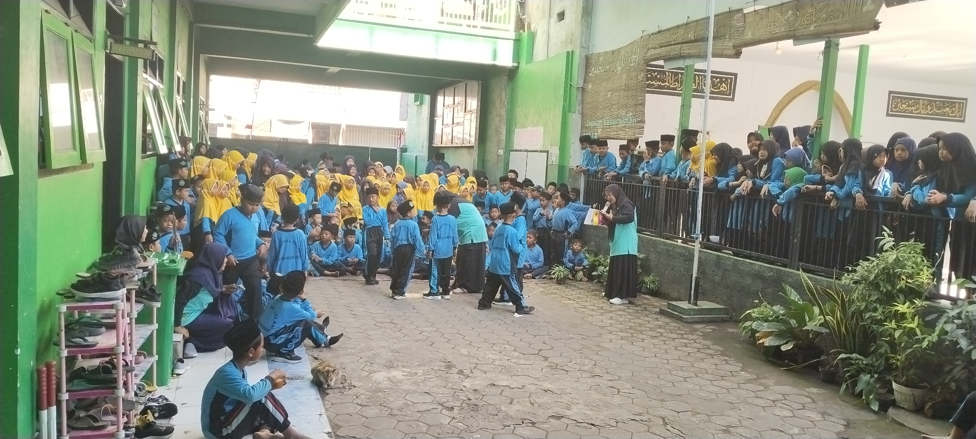 The image depicts an outdoor scene in front of a building with green walls, possibly a school in Yosowilangun, Indonesia. A large group of children, predominantly in blue uniforms and some in yellow headscarves, are gathered. Some are standing, while others are sitting on a paved courtyard. Several adults, including a woman speaking into a microphone, are also present. In the foreground, there's a shoe rack filled with footwear and a cat lying on the ground. The background shows a covered walkway and some Arabic calligraphy on the wall. The overall atmosphere appears to be an event or gathering, likely a school activity.