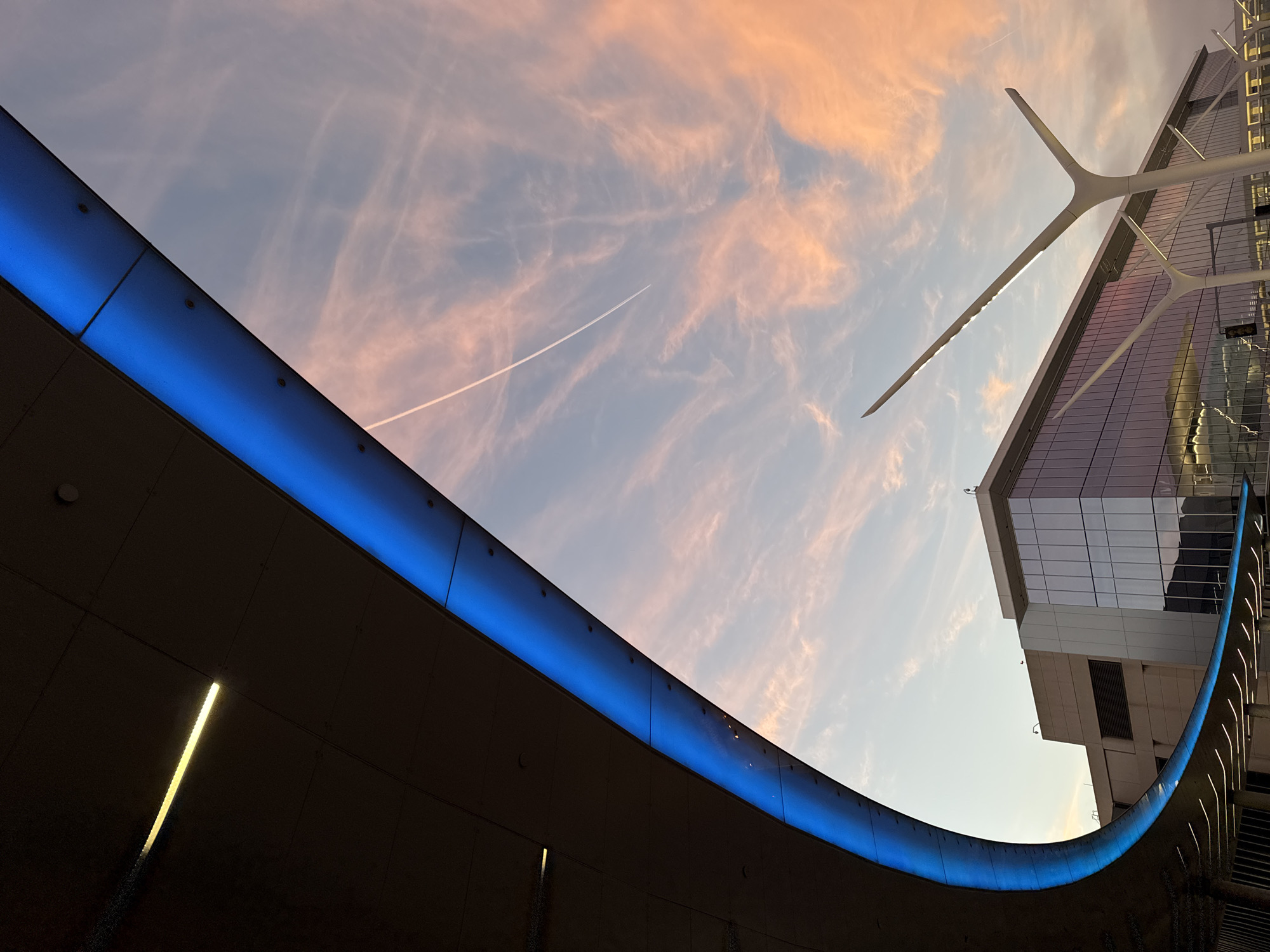 The image portrays an exterior architectural view at Los Angeles International Airport (LAX) in Los Angeles, California, United States, during twilight conditions. The upper two-thirds of the frame show a sky with scattered, wispy clouds illuminated in shades of pink and orange, transitioning to a pale blue-grey. A faint white aircraft contrail extends diagonally from the upper left to the middle right portion of the sky.
Dominating the upper left and curving towards the lower center is a large, dark-toned overhead structure, identified as part of the People Mover guideway. Its underside features a continuous strip of illuminated blue LED lighting along its curved edge. Further back along the darker underside of this structure, a horizontal yellow linear light is visible. In the mid-ground, below the overhead structure and against the horizon, stands a modern airport terminal building characterized by large, reflective glass curtain walls and sections of light-colored paneling. To the lower right, a distinctive white, multi-branched "pylon tree" streetlight, characteristic of LAX's design, extends upwards, with its integrated light source emitting illumination. No discernible individuals or active vehicles are present in the visible area.