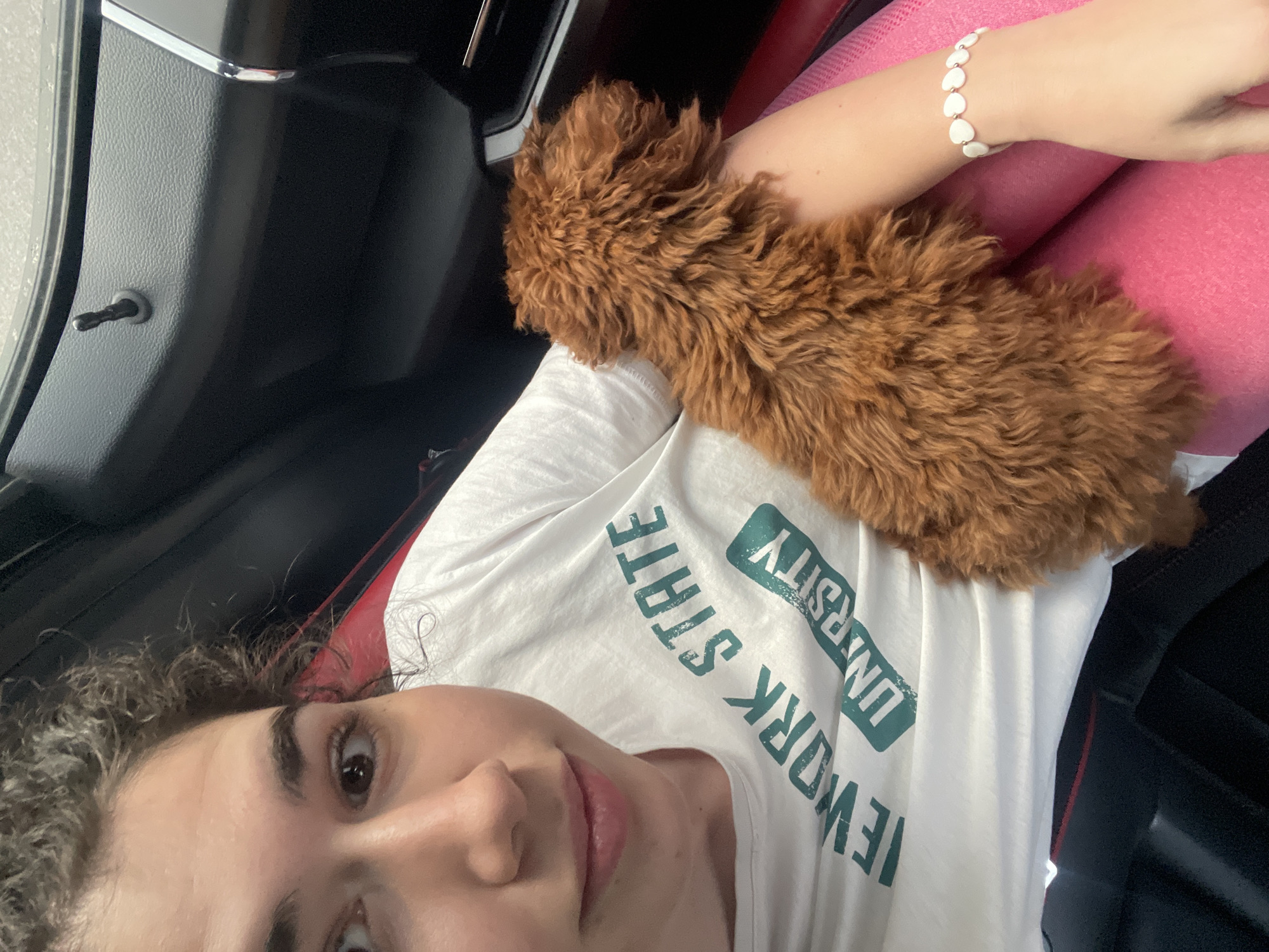 A young woman is captured in a selfie-style portrait, positioned inside a car, likely in the passenger seat. She has curly dark hair and dark eyes, looking directly at the camera with a gentle, relaxed expression and a slight smile. She is wearing a white t-shirt with green text that reads "NEW YORK STATE UNIVERSITY" and pink pants or leggings. On her right wrist, she wears a bracelet made of white, heart-shaped beads.
Resting comfortably on her arm and lap is a small, fluffy, brown dog, possibly a puppy of a poodle or doodle breed, its soft, curly fur prominent in the foreground. The dog appears calm and content.
The interior of the car is visible, featuring a black door panel to the left with a chrome handle and a visible door lock pin, confirming the setting is inside a vehicle. The lighting suggests it is daytime. The overall scene depicts a casual and affectionate moment between the woman and her dog. Based on the provided context, the location is Sofia, Bulgaria, although no specific visual cues in the image confirm this.