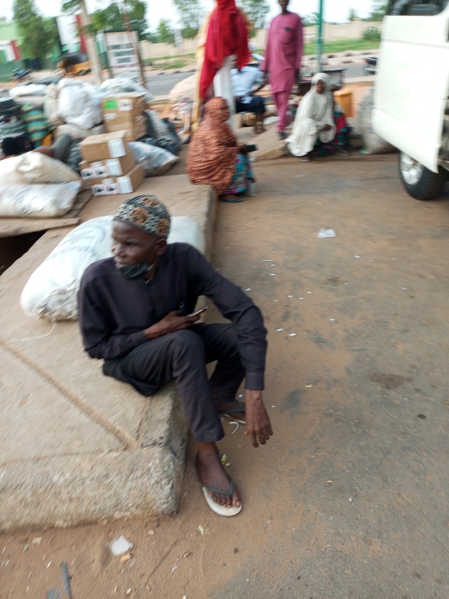 A man wearing a dark long-sleeved shirt, dark trousers, and a patterned cap is sitting on a concrete ledge. He is holding a mobile phone in his right hand and appears to be looking to his right. A black face mask is pulled down below his chin. Beside him, to his left, are several large white sacks. In the background, other people are visible, including a woman in a red hijab and a man in a pink outfit. Various bags, boxes, and a white vehicle are also present. The scene is set on a dusty outdoor surface, likely a market or roadside area in Gombe, Nigeria, given the context of the request.