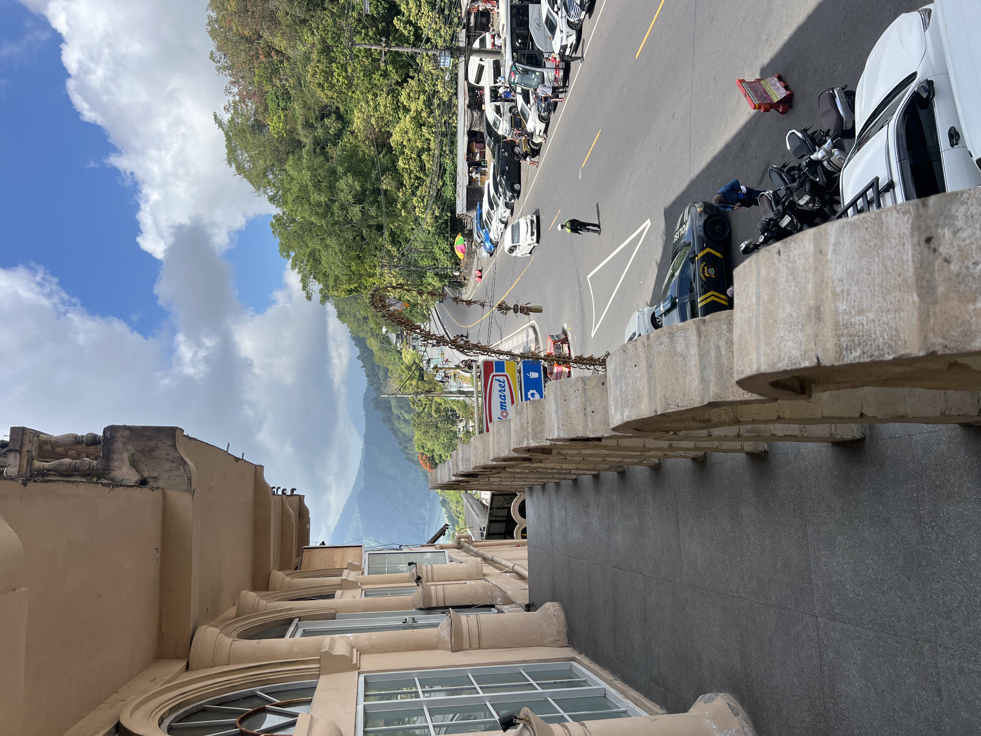 This is a street-level view of a bustling road in Kintamani, Indonesia, taken from an elevated vantage point. The foreground features a stone balustrade and a dark grey tiled walkway, part of a building with arched windows and a beige facade.
The road itself is paved and curves upwards into a lush, green hillside. It is lined with parked cars and motorcycles, suggesting a popular tourist area. Several people are visible, including a person in a dark uniform standing near a police car and another individual walking on the road. A small market or food stall area is set up along the side of the road, indicated by corrugated metal roofs and people gathered around.
A prominent "Indomaret" sign is visible, a common convenience store chain in Indonesia. The backdrop consists of rolling hills and distant mountains, with a cloudy blue sky overhead. The overall scene depicts a lively, everyday environment in a scenic, mountainous region.