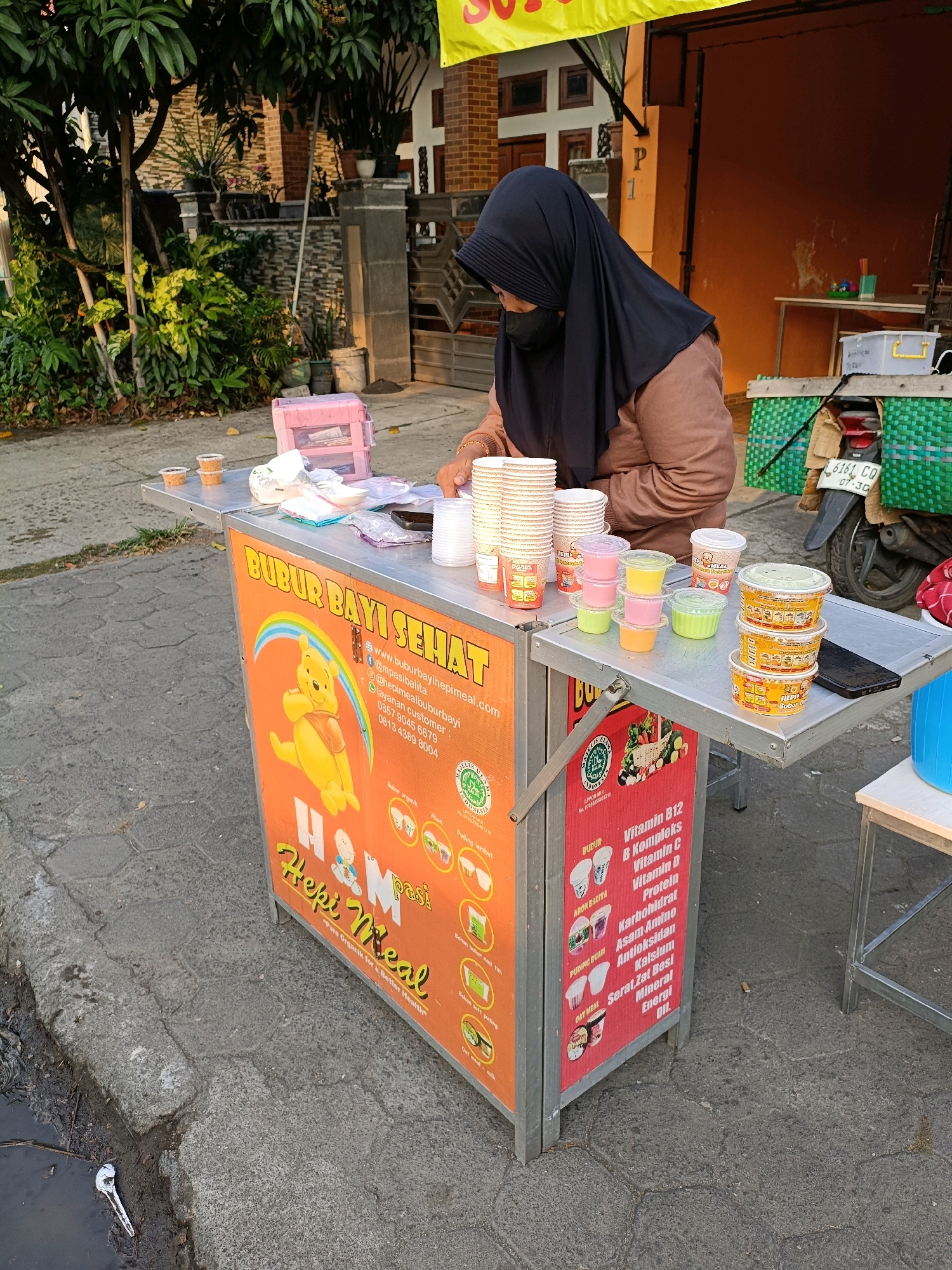 The image depicts an outdoor street stall in Purwomartani, Indonesia, selling baby food. A woman wearing a black hijab and brown top is standing behind the stall, preparing or serving food. The stall itself is an orange cart with a foldable metal table on the right side. The cart is adorned with colorful graphics, including cartoon characters and product information in Indonesian. Various food items are displayed on the table, including stacks of small cups filled with colorful baby food, larger containers of baby food, and piles of disposable cups. A motorcycle with a green woven basket is parked behind the stall. The background shows a brick building, lush green trees, and a paved street. The scene appears to be captured in the late afternoon or early morning, with the sunlight casting shadows.