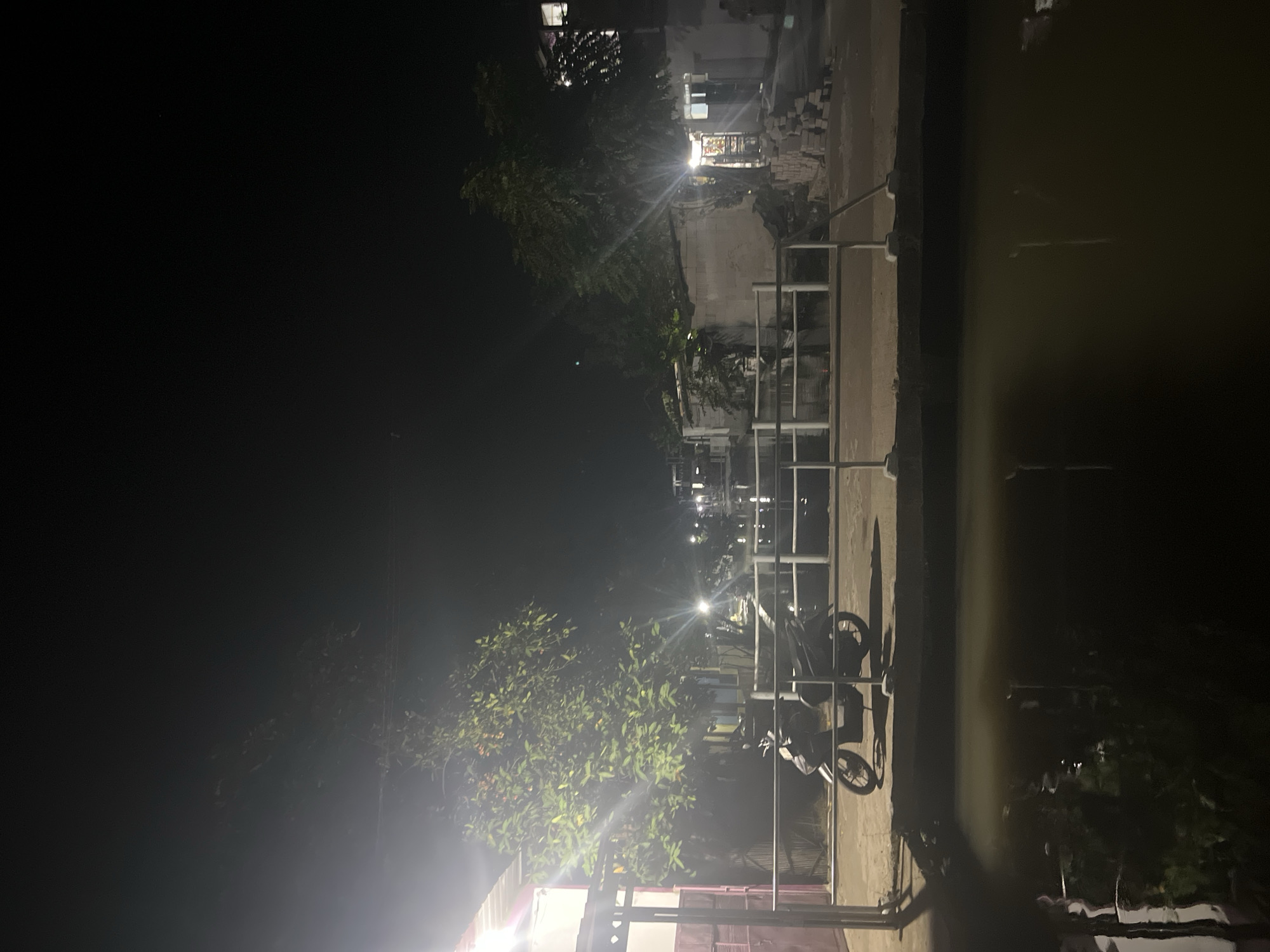 The image depicts a dimly lit street scene at night in Laban, Indonesia. In the foreground, a body of water reflects the ambient light and surrounding structures. A concrete walkway with a white railing runs along the edge of the water, and a parked scooter is visible on the walkway. Beyond the walkway, there are several buildings, some appearing to be houses or small shops, illuminated by artificial lights. Trees with green foliage are scattered throughout the scene, their leaves catching the light. The overall impression is of a quiet residential or commercial area at night, with a mix of man-made structures and natural elements.