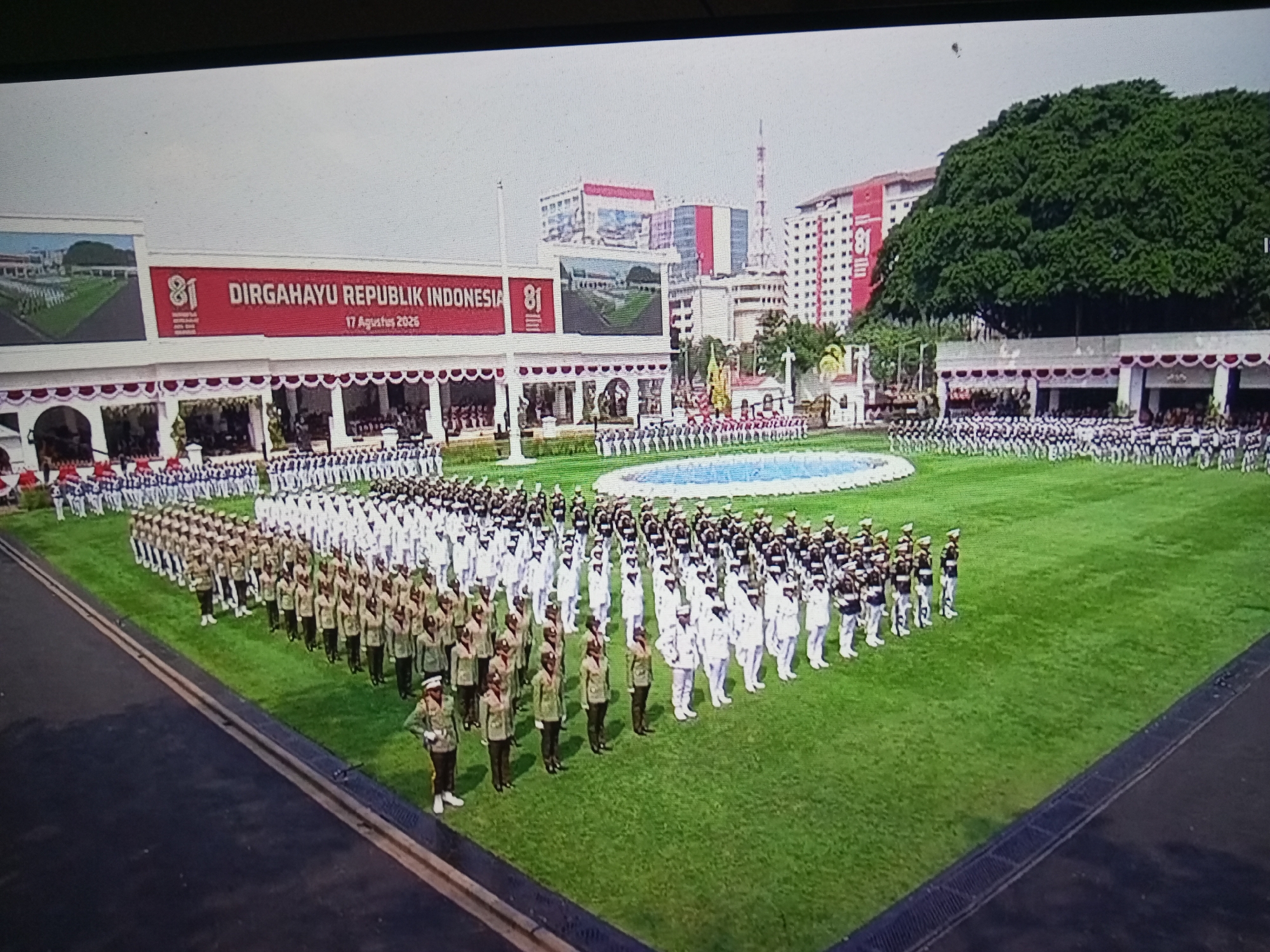 The image captures a large-scale ceremonial event on a bright, overcast day, likely in Banyudono, Indonesia, judging by the "DIRGAHAYU REPUBLIK INDONESIA" banner celebrating Indonesia's 81st Independence Day on August 17, 2026. The foreground shows a manicured green field with several rows of uniformed individuals standing at attention. Closest to the viewer are individuals in light green uniforms with darker green details and hats. Behind them are rows of people in white uniforms and dark trousers, some with hats. Further back, more uniformed groups are visible, some in white and dark uniforms, suggesting a diverse representation of military or official organizations.
The middle ground features a large, white, open-air pavilion-like structure adorned with red and white banners, a large screen displaying an image of a ceremonial gathering, and the prominent "81 DIRGAHAYU REPUBLIK INDONESIA 17 Agustus 2026" text. Behind this structure, modern buildings rise, including a red and white building with "81" prominently displayed. A very large, dense green tree dominates the upper right portion of the image, providing a stark contrast to the urban architecture. The sky is a pale, washed-out grey, typical of an overcast day. The overall scene depicts a formal national celebration, likely an independence day parade or ceremony, with a focus on uniformed personnel and patriotic displays.
