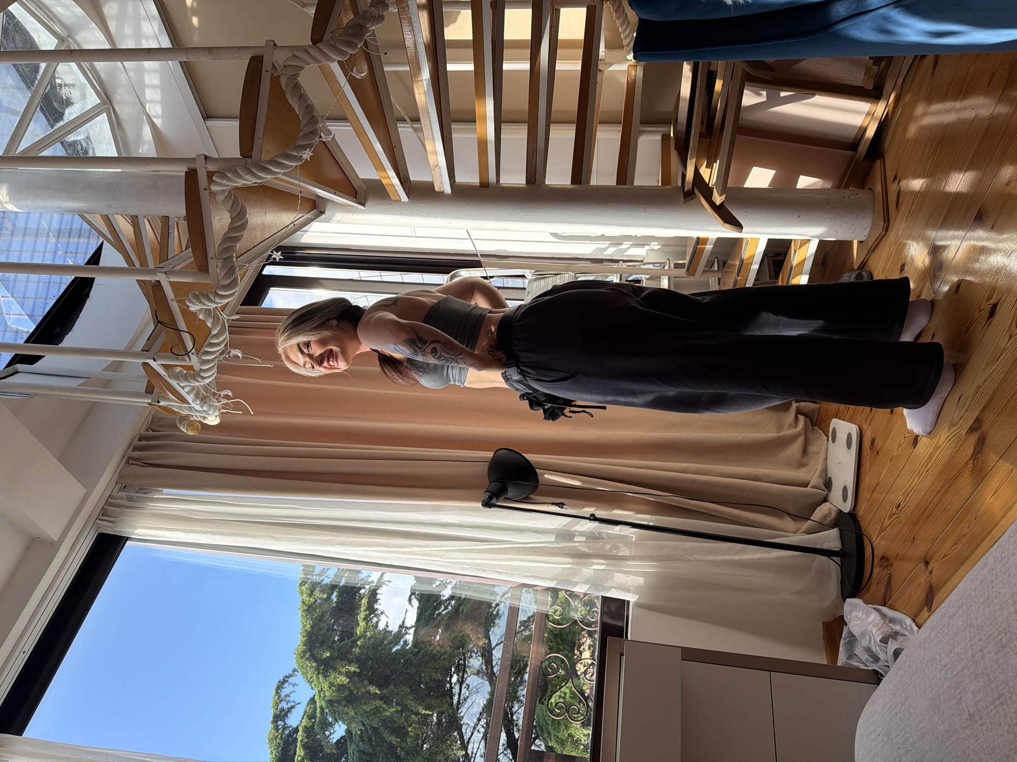 The image shows a woman in a gray sports bra and dark sweatpants, standing indoors. She is smiling and looking towards the camera. She has tattoos on her arms. The setting is a room with a spiral staircase, wooden floors, and large windows that reveal a view of greenery and clear blue sky. A floor lamp is visible near the window, and a digital scale sits on the wooden floor. The location is likely in Budva, Montenegro, given the style of architecture and lush Mediterranean vegetation visible through the window.