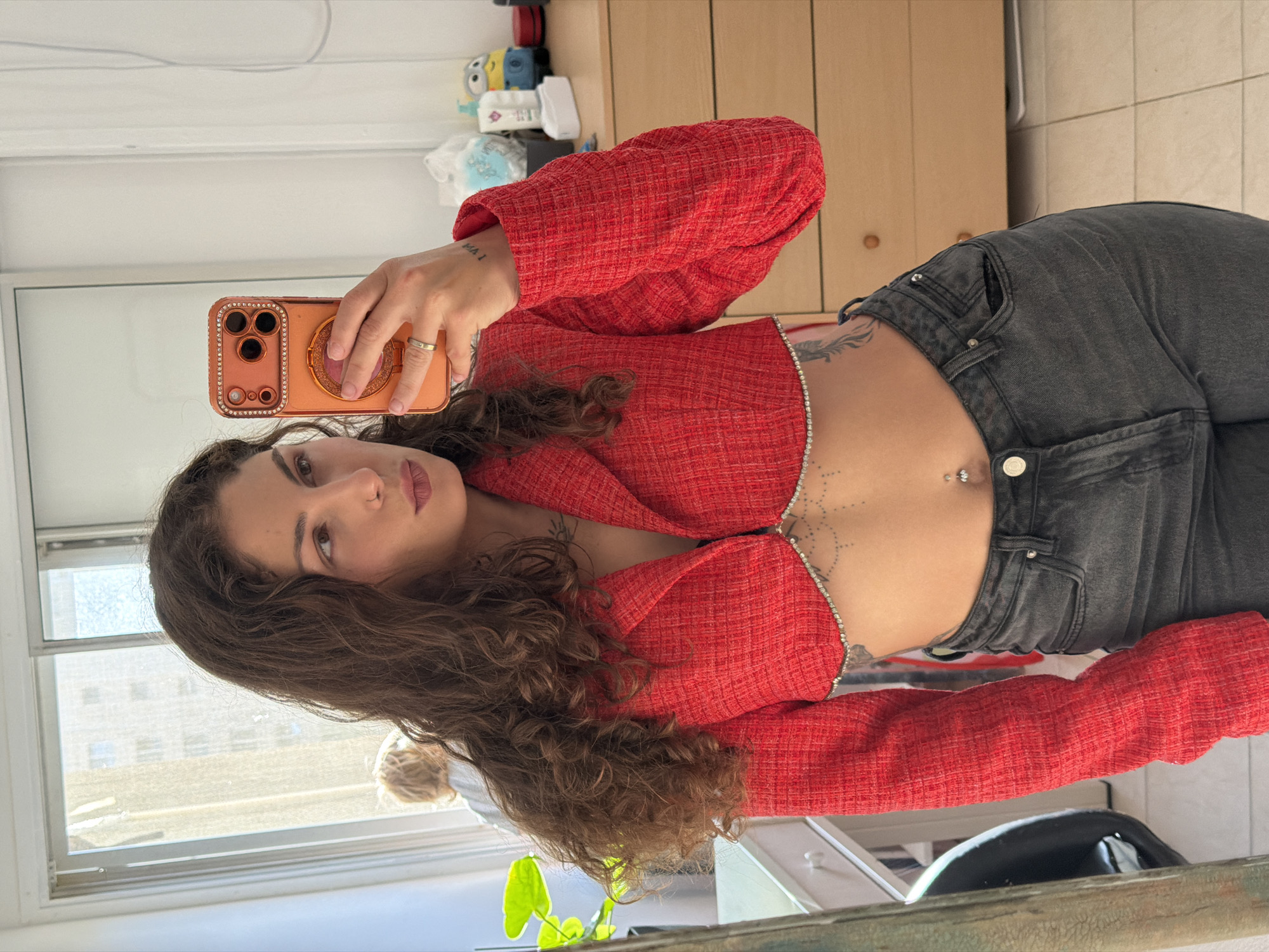 A young woman with long, curly brown hair is taking a selfie using an orange smartphone with a jeweled case. She's wearing a red, cropped jacket with long sleeves, paired with dark gray jeans. A decorative chain adorns the front of her torso, and she has a piercing above her navel. In the background, a window reveals a cityscape, suggesting an urban environment. An indoor cabinet and household objects are also visible, indicating the picture was taken indoors. There is no indication this photo was taken in Kiryat Bialik. More information is needed to provide an assessment regarding the location.