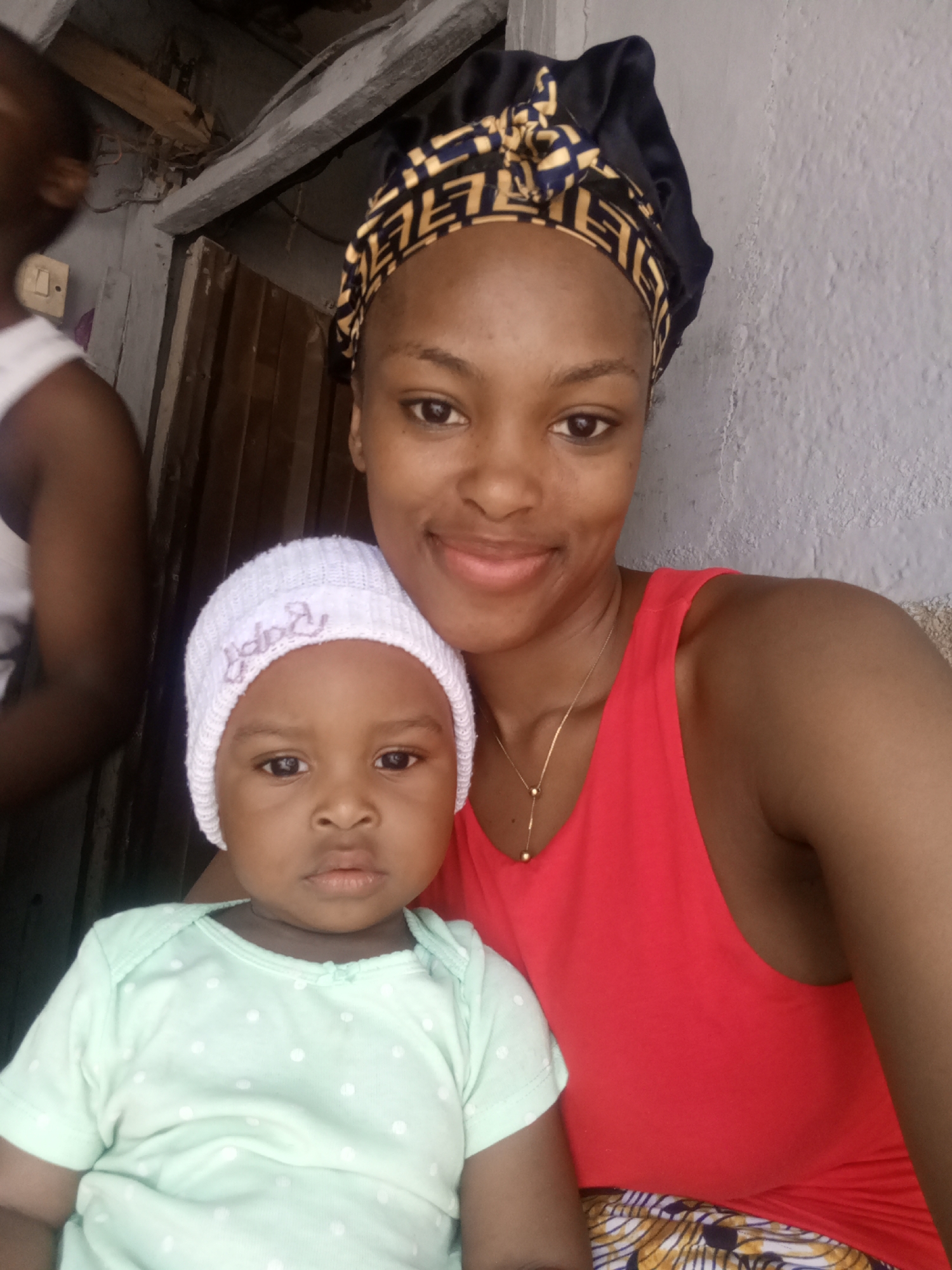 A medium shot shows two subjects, a mother and her baby, against a muted background. The mother, on the right, wears a black and gold patterned headscarf and a bright red sleeveless top. She has a gold necklace with two spherical pendants. The baby, on the left, is wearing a white beanie with "Baby" embroidered in pink thread and a light green polka-dotted onesie. They are in an outdoor or semi-outdoor setting, with a grey wall to the right and a wooden structure or door to the left, suggesting an urban or semi-urban environment, possibly in a residential area. The lighting is natural and somewhat diffused. The composition is a selfie, with the mother framing the shot.