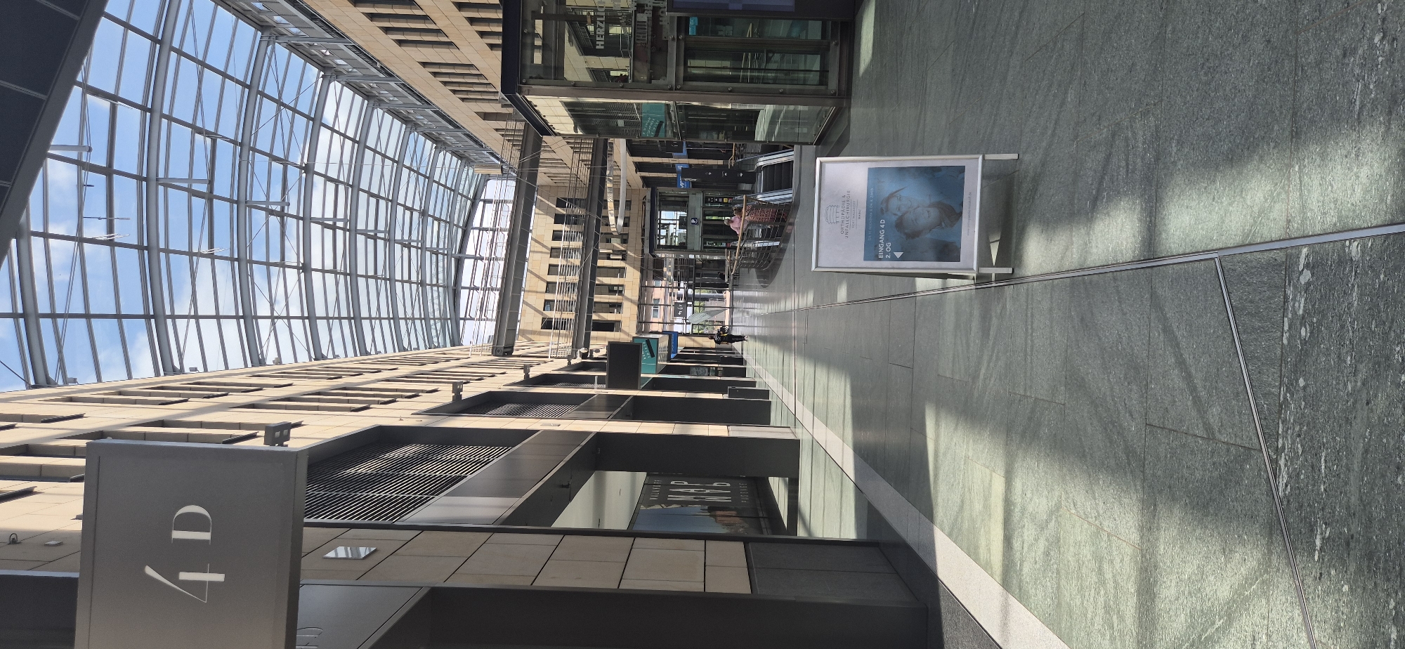 This image depicts the interior of a modern, multi-story building, likely a shopping arcade or office complex, with a curved glass ceiling that allows natural light to filter in. The architecture features clean lines, stone flooring, and tan-colored facades of the buildings lining the walkway. On the left, a sign reads "4D".
In the foreground, an A-frame sign displays an advertisement for "ORTHOPÄDIE & UNFALLCHIRURGIE" (Orthopedics & Trauma Surgery) and "EINGANG 4D 2. OG" (Entrance 4D, 2nd Floor). The sign also features an image of two individuals.
Further down the walkway, several people are visible. One individual walks towards the camera, while two others are near escalators on the right side of the image. The environment is well-lit, with sunlight casting distinct shadows on the floor, suggesting it is daytime.
The building's facade is characterized by repetitive windows and metal walkways connecting different levels. The overall impression is one of a contemporary, spacious, and well-maintained indoor public space.
This location is identified as the "Forum Gutenberg" in Mainz, Germany, which houses various businesses and medical practices, including the orthopedics and trauma surgery clinic advertised on the sign.