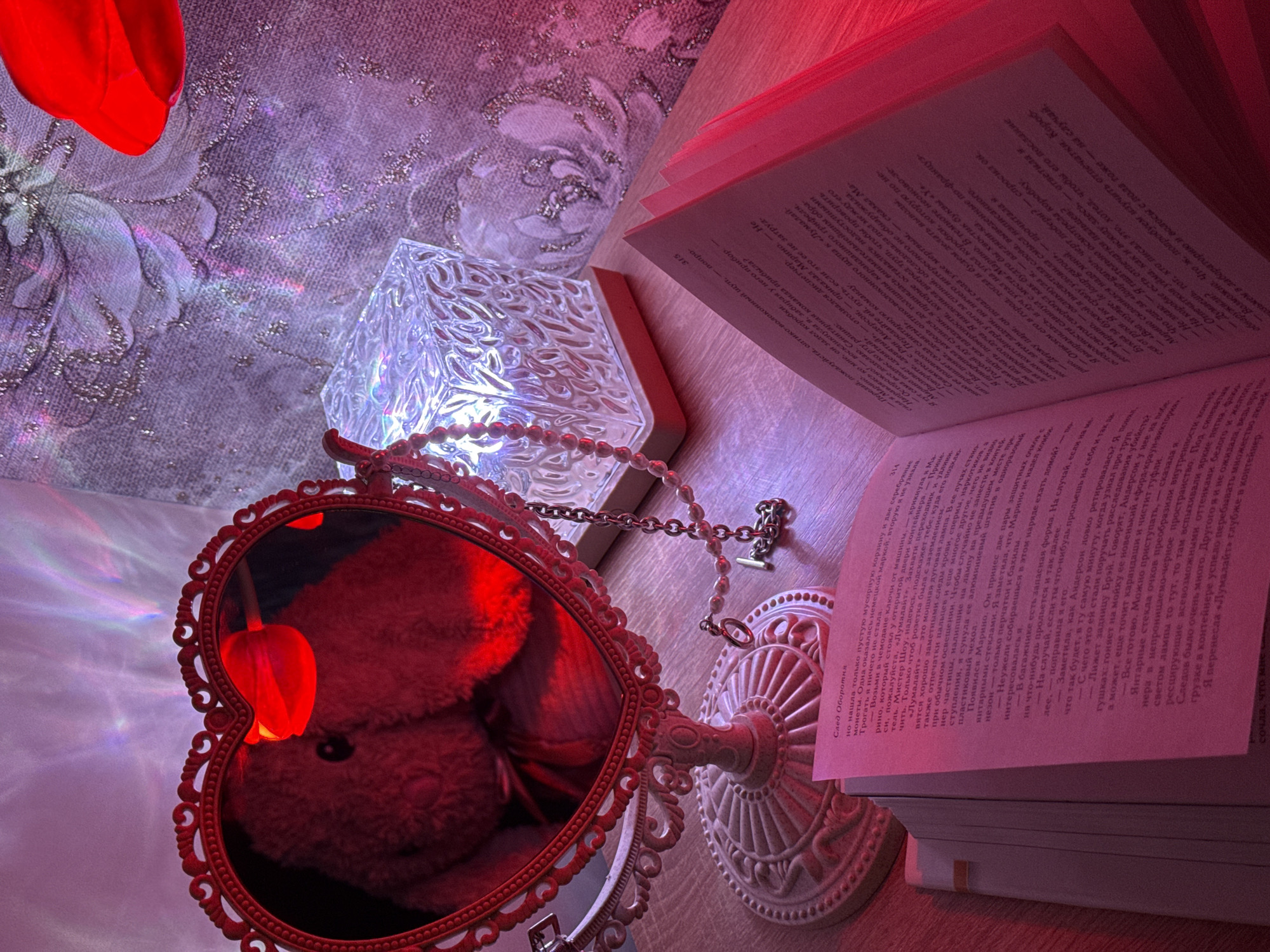The image depicts a still life composition on a wooden surface, possibly a table, under predominantly red and purple lighting. The scene includes a partly open book with Russian text, a heart-shaped ornate mirror reflecting a blurred plush toy, a decorative translucent cube emitting light, and a string of decorative beads. The background features a textured surface with floral patterns. The overall atmosphere suggests a cozy or romantic setting, though the exact location is indeterminate.