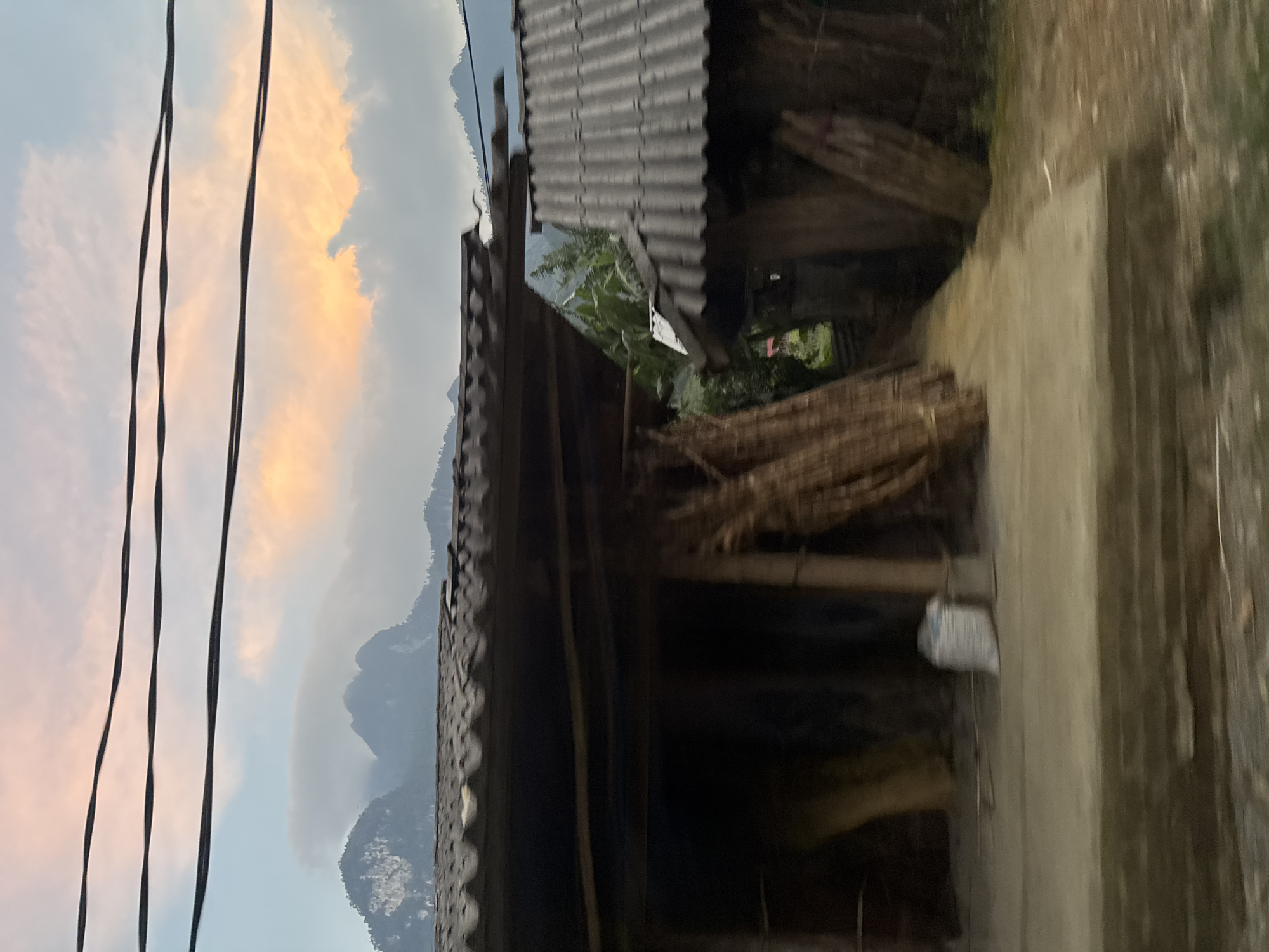 The image shows a view in Du Già Commune, Vietnam. The scene includes mountains in the background, visible through the sky with clouds tinted with orange. Power lines are visible against the sky. A corrugated metal roof covers a structure to the right of the frame. Inside the structure, bundles of sticks or bamboo are stored. A concrete surface is partially visible to the right, with a white object hanging or resting in the shadowed space. The photograph is slightly blurred and rotated counter-clockwise.