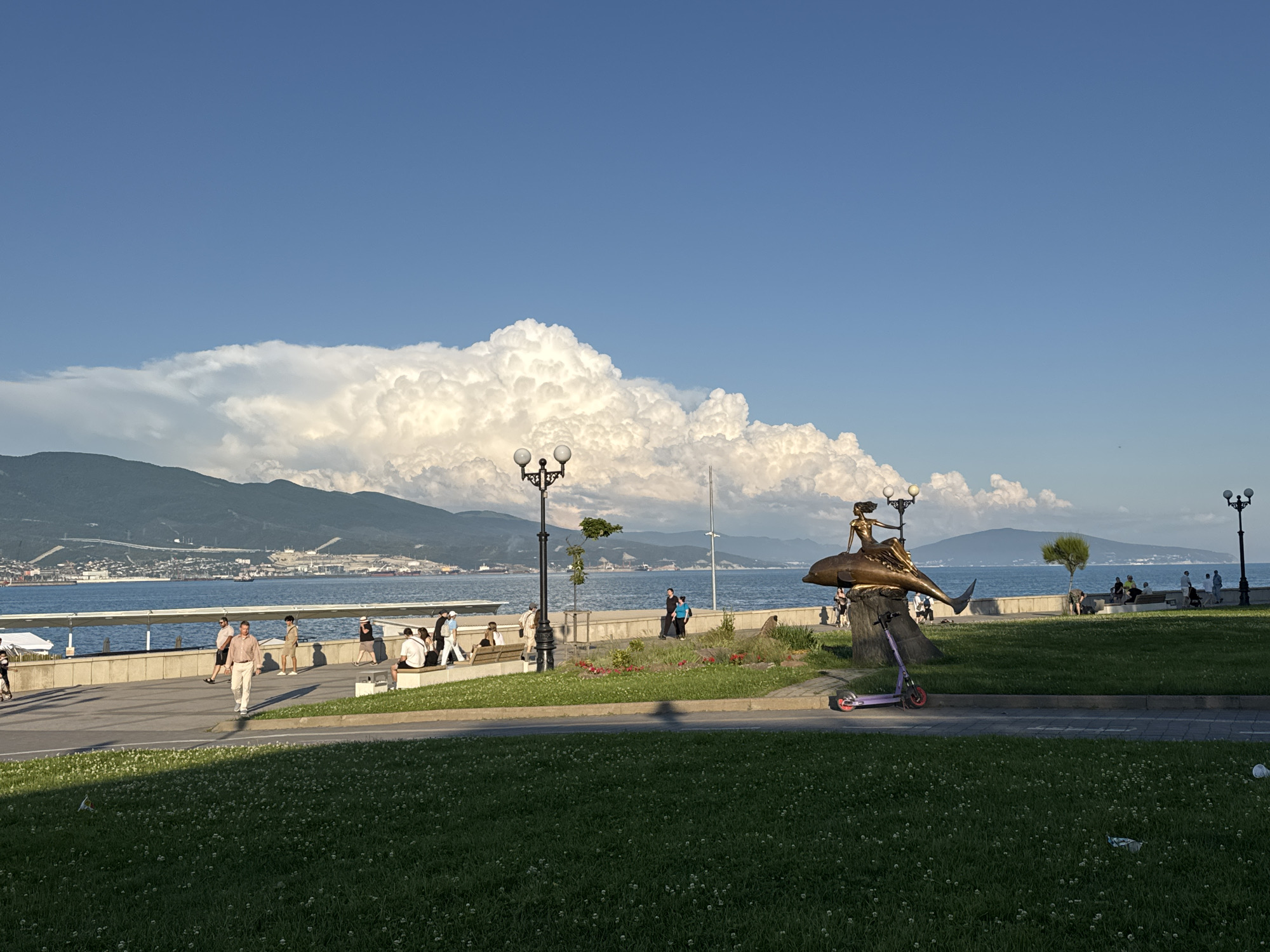 The image depicts a scenic waterfront promenade with a clear blue sky and dramatic, large cumulus clouds forming over distant mountains. In the foreground, a well-maintained grassy area is visible, dotted with small white flowers. A paved walkway runs parallel to the water, separating the grassy area from a concrete embankment.
Along the promenade, several people are casually strolling and sitting on benches. To the right of the image, a bronze sculpture of a woman riding a dolphin is prominently displayed. A purple electric scooter is parked in front of the sculpture. The sea stretches out beyond the embankment, with several ships and boats visible on the horizon. The distant shoreline is lined with buildings and industrial structures, suggesting a port or coastal city. The overall atmosphere is one of a pleasant, sunny day by the sea. The scene is likely located in Grushovaya Balka, Russia, given the context of the request.