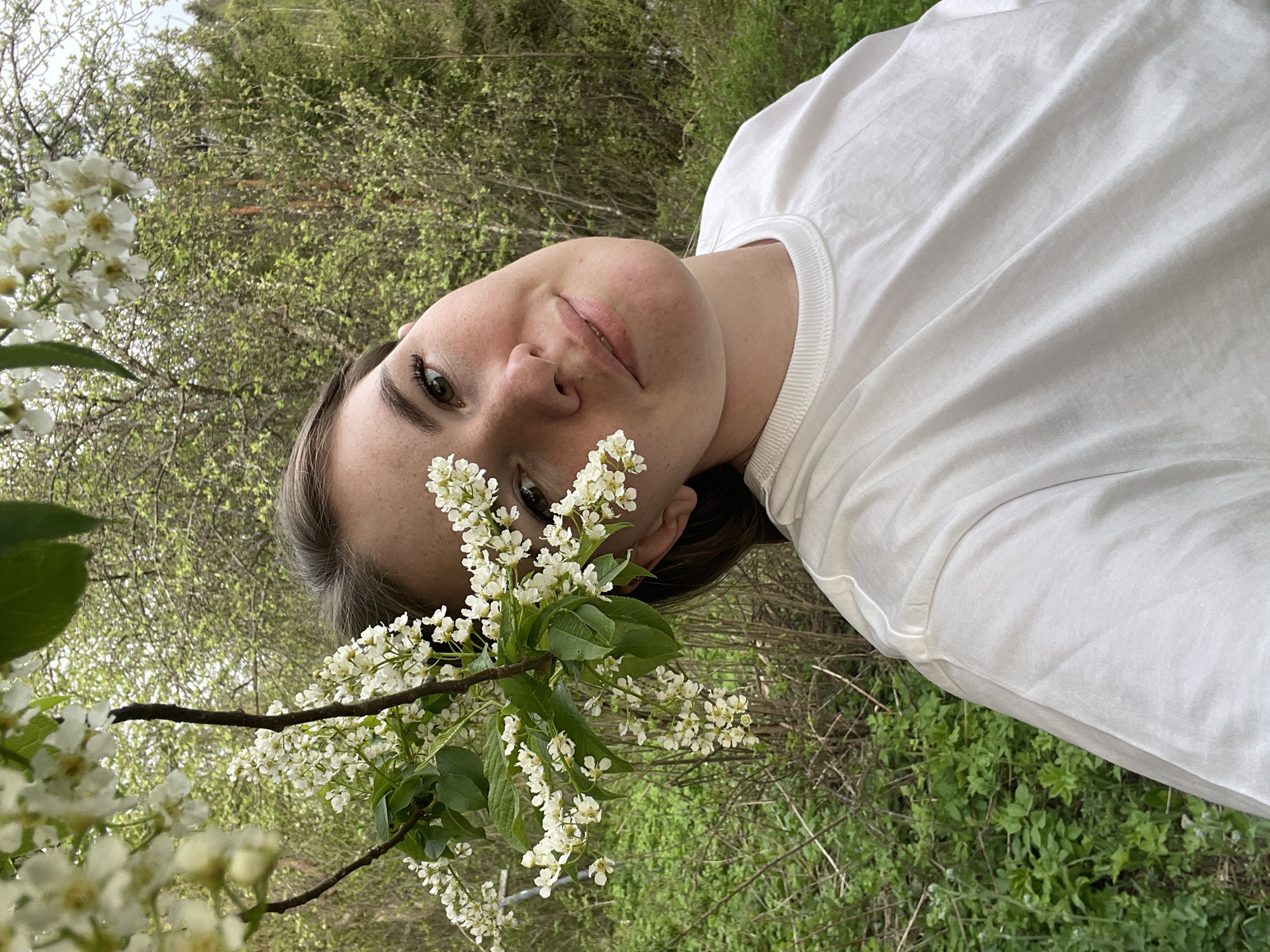A young woman with fair skin is lying on her side outdoors, with her head near a flowering tree branch. She is wearing a white T-shirt. The background includes greenery, suggesting a natural environment such as a forest or park. The location is in Zhigalki, Russia.