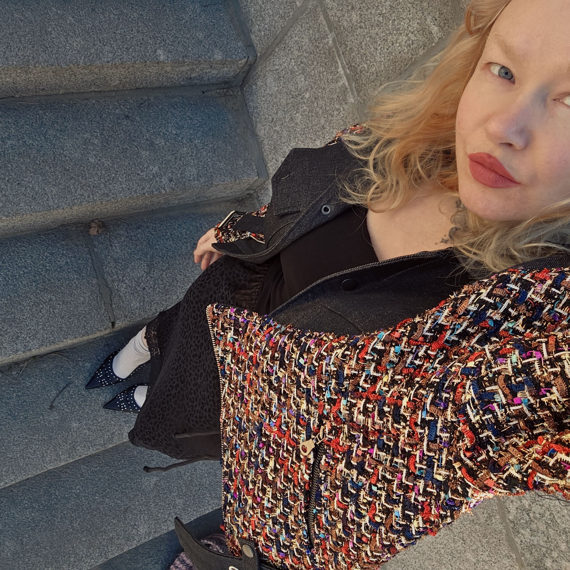 A woman with blonde wavy hair and blue eyes sits on grey stone steps, likely in Saint Petersburg, Russia, based on the architectural style. She wears a black top, a tweed-like jacket with colorful threads, a black jacket, and a printed skirt. Her feet are clad in white socks and pointed-toe shoes adorned with studs. A tattoo is visible on her neck. The photograph is taken from an elevated perspective.