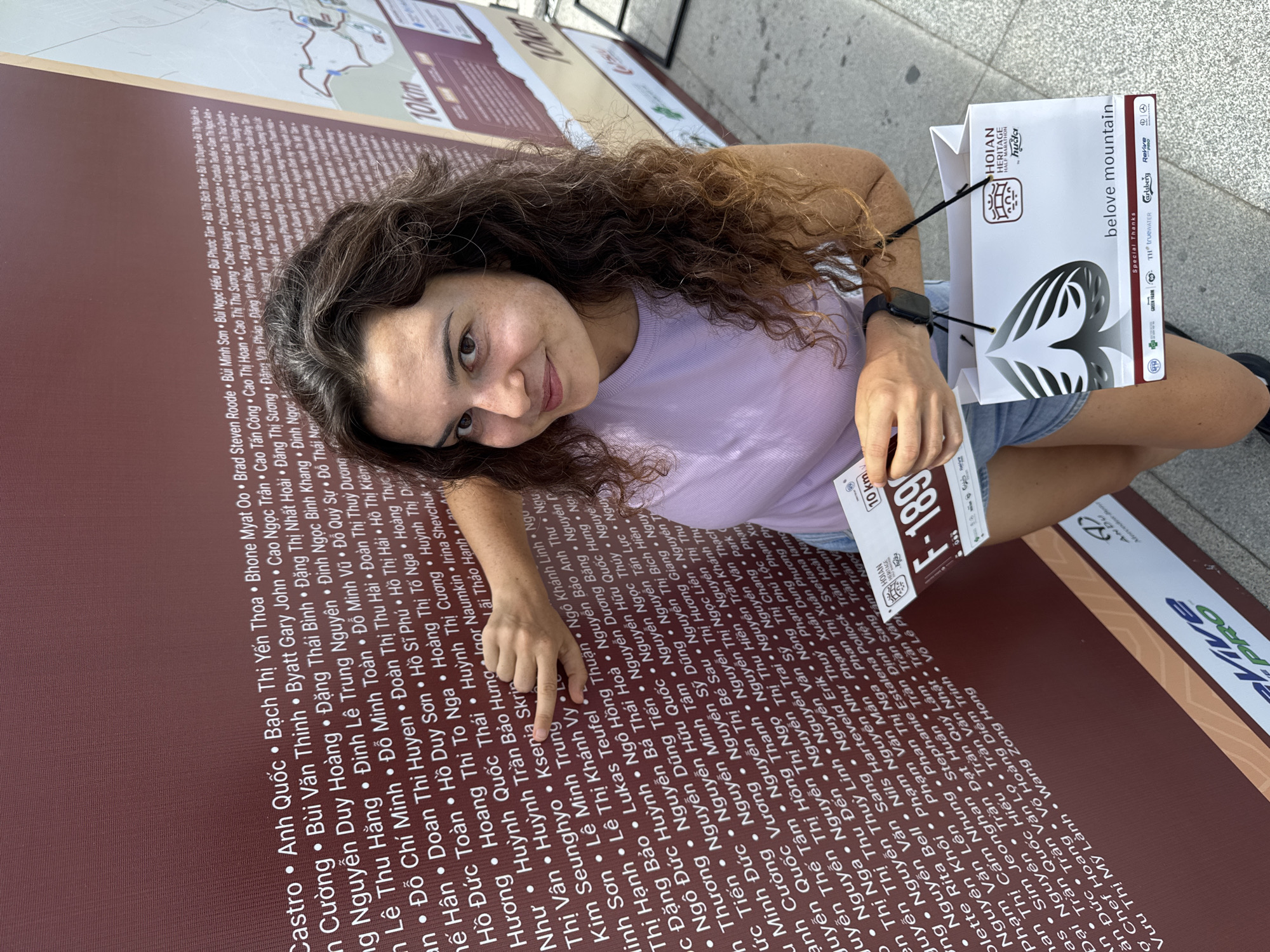 A young woman with curly brown hair and fair skin, wearing a light purple t-shirt and denim shorts, stands in front of a large, brown wall with a list of names printed in white text. She is smiling and holding a small white card with the text "F-189" and "10 km" visible. In her other hand, she holds a white shopping bag with black handles, which has the text "HOIAN HERITAGE HALF MARATHON" and "belove mountain" printed on it. The wall appears to be a display related to the marathon. The ground is paved with light gray concrete. In the background, a map of the Hoi An area with a 10km marker is visible.