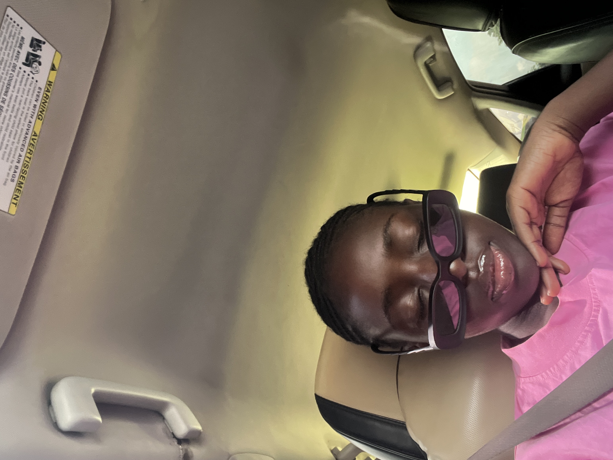 A dark-skinned female with braided hair and sunglasses rests her chin on her hand while sitting in the passenger seat of a car. She is wearing a bright pink t-shirt. The interior of the car is visible, including the beige headrest of the passenger seat and the black leather headrest of the driver's seat. A yellow safety warning sticker is affixed to the car's ceiling. The background is blurred, but the sky and trees are visible through the car window, suggesting an outdoor location. The scene appears to be in Kuchingoro, Nigeria.