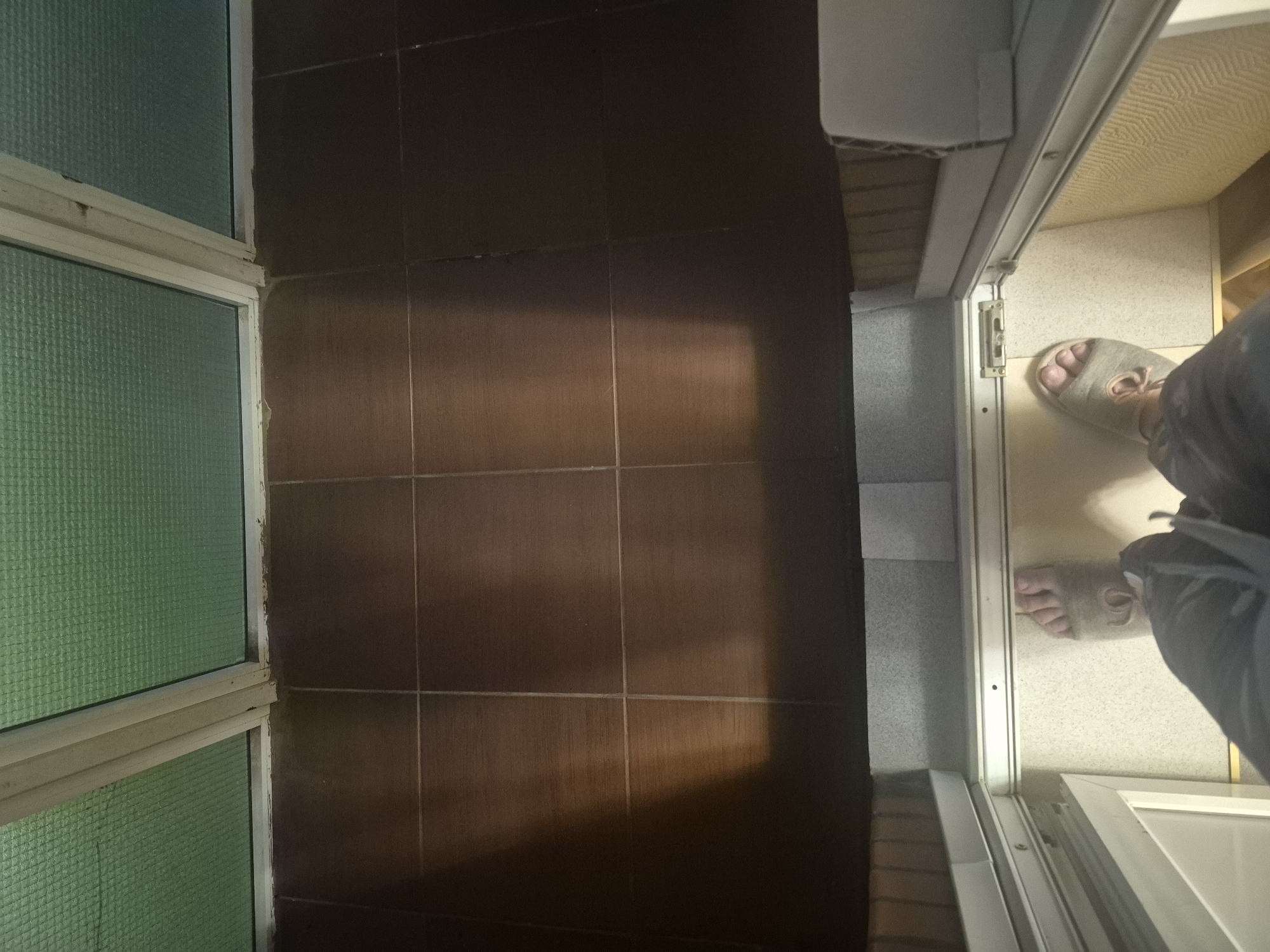 The image depicts a view from inside a room looking out through a partially open door. The scene is a domestic interior, likely a bathroom or hallway, characterized by dark brown tiled walls and a lighter tiled floor. A section of a window with a textured green glass is visible on the left, suggesting a bathroom or a room with a window facing an exterior that receives sunlight.
The primary subjects are a person's feet wearing light grey slippers, visible through the open doorway. The person is standing on the lighter tiled floor. The angle of the image, taken from a low perspective, emphasizes the floor and the lower extremities of the person. The lighting suggests it is daytime, with a distinct shadow cast across the tiled wall, indicating sunlight entering from the open door.
The doorway itself appears to be a sliding door with a white frame. A locking mechanism is visible on the door frame. The scene is simple and utilitarian, with no apparent decorative elements. The overall impression is one of everyday domestic life, possibly someone entering or leaving a room.
The location context of Saint Petersburg, Russia, does not offer specific visual cues within the image itself, but it provides a general geographical setting. The weather cannot be determined from the image. There is no visible text in the image.