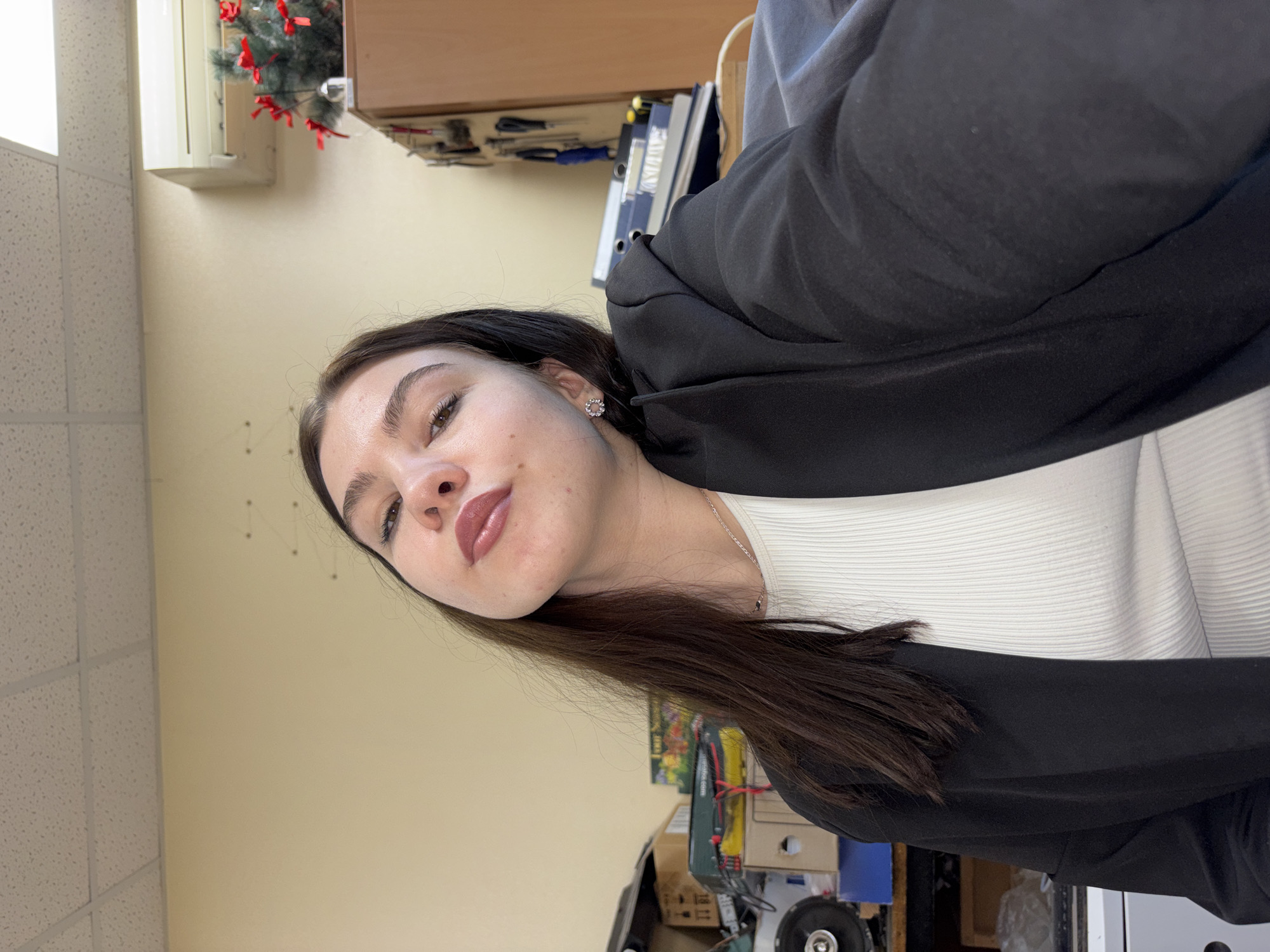 A photograph shows a woman with dark hair and fair skin, wearing a white top and black jacket. She is indoors, likely in an office or workplace setting, based on the presence of ceiling tiles and office equipment in the background. The location is in Perm, Russia. Visible in the background are a window with a small Christmas decoration, files, boxes, and various office supplies. The woman is oriented at an angle to the camera, looking directly at the viewer.