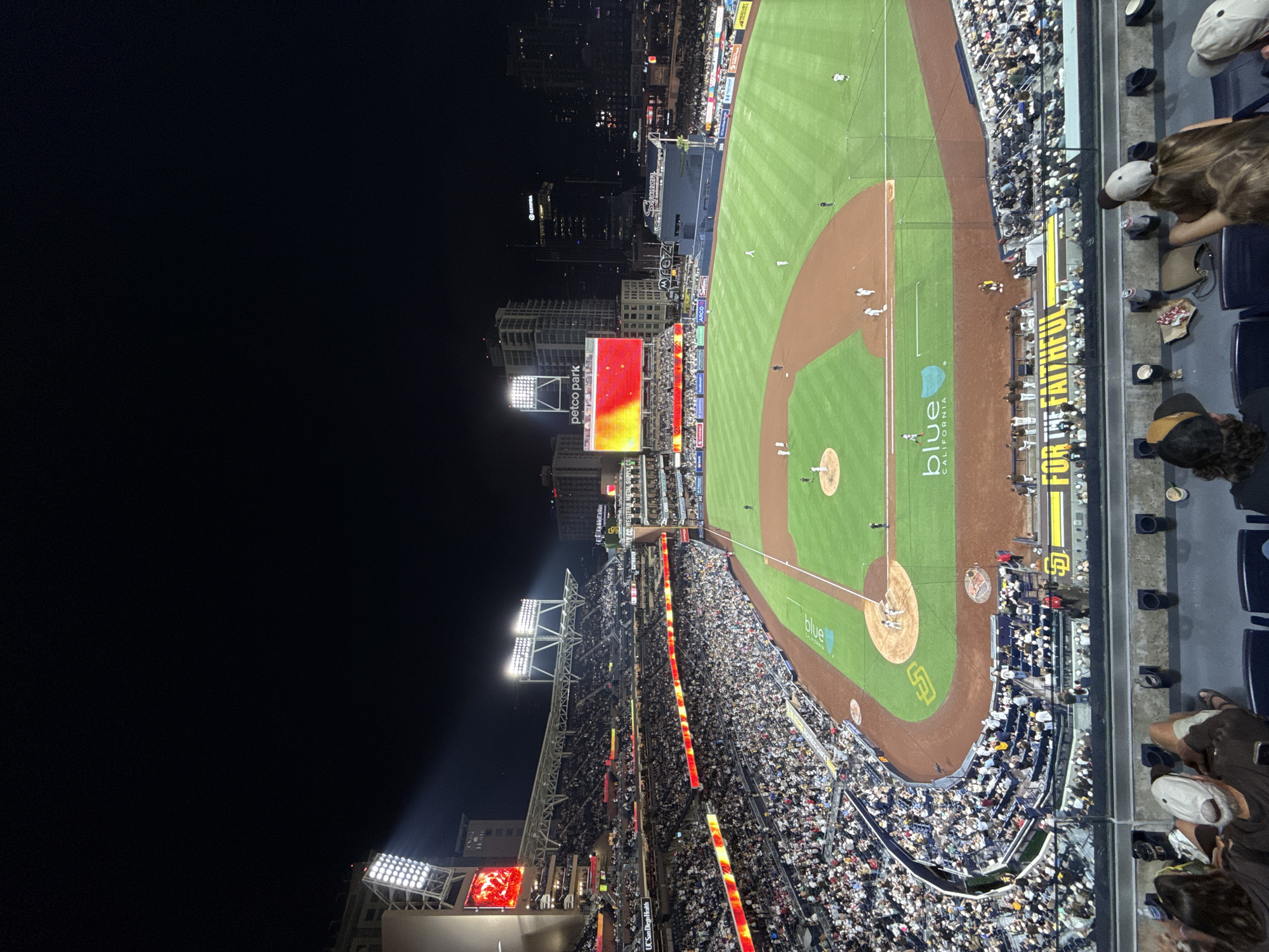 This aerial, nighttime photo shows a bustling baseball stadium, likely Petco Park in San Diego, California, based on the visible text. The field is illuminated, showcasing a baseball game in progress with players on the field. Stadium lights are visible, casting beams across the green turf and brown dirt infield. A large scoreboard displays a vibrant, fiery image.
Thousands of spectators fill the tiered seating surrounding the field, with many visible in the foreground and along the sidelines. The crowd appears engaged, suggesting an exciting game. Some fans in the foreground are partially visible, wearing caps and looking towards the game. The city skyline with lit buildings can be seen in the background, indicating an urban setting at night.
Prominent text on the scoreboard reads "petco park," confirming the location. Other visible text includes "UC San Diego Health," "PLY CO.," "blue CALIFORNIA," and "FOR THE FAITHFUL," which is emblazoned on a yellow banner. The overall atmosphere appears energetic and lively, characteristic of a major sporting event. The time of day is clearly night, with the stadium lights and city lights being the primary sources of illumination.