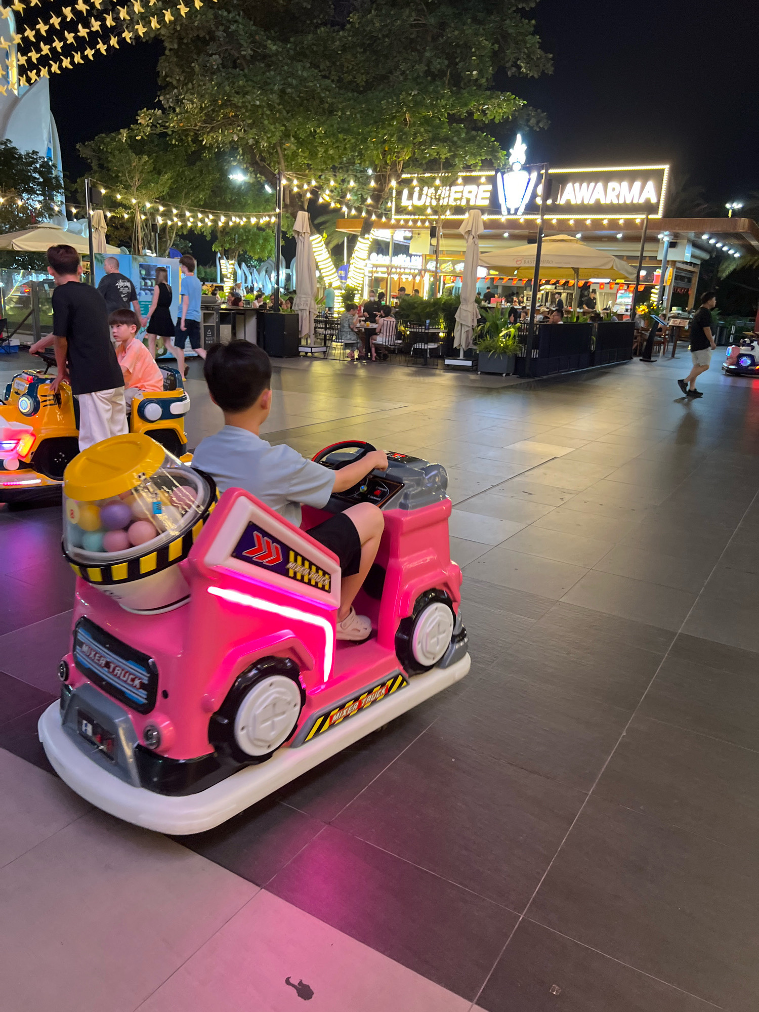 In Nha Trang, Vietnam, a young boy is seated in a pink toy mixer truck ride in an outdoor entertainment area at night. The truck has lights and a transparent container filled with colorful balls on its side. The boy is wearing a light blue t-shirt and dark shorts, and he is holding the steering wheel. Behind him, another boy in an orange t-shirt sits in a yellow toy truck. Several other people are visible in the background, including adults and children, walking and seated at tables in a restaurant area. String lights are strung overhead, illuminating the scene. A sign in the background reads "LUMIERE Shawarma". The ground is paved with dark tiles.
