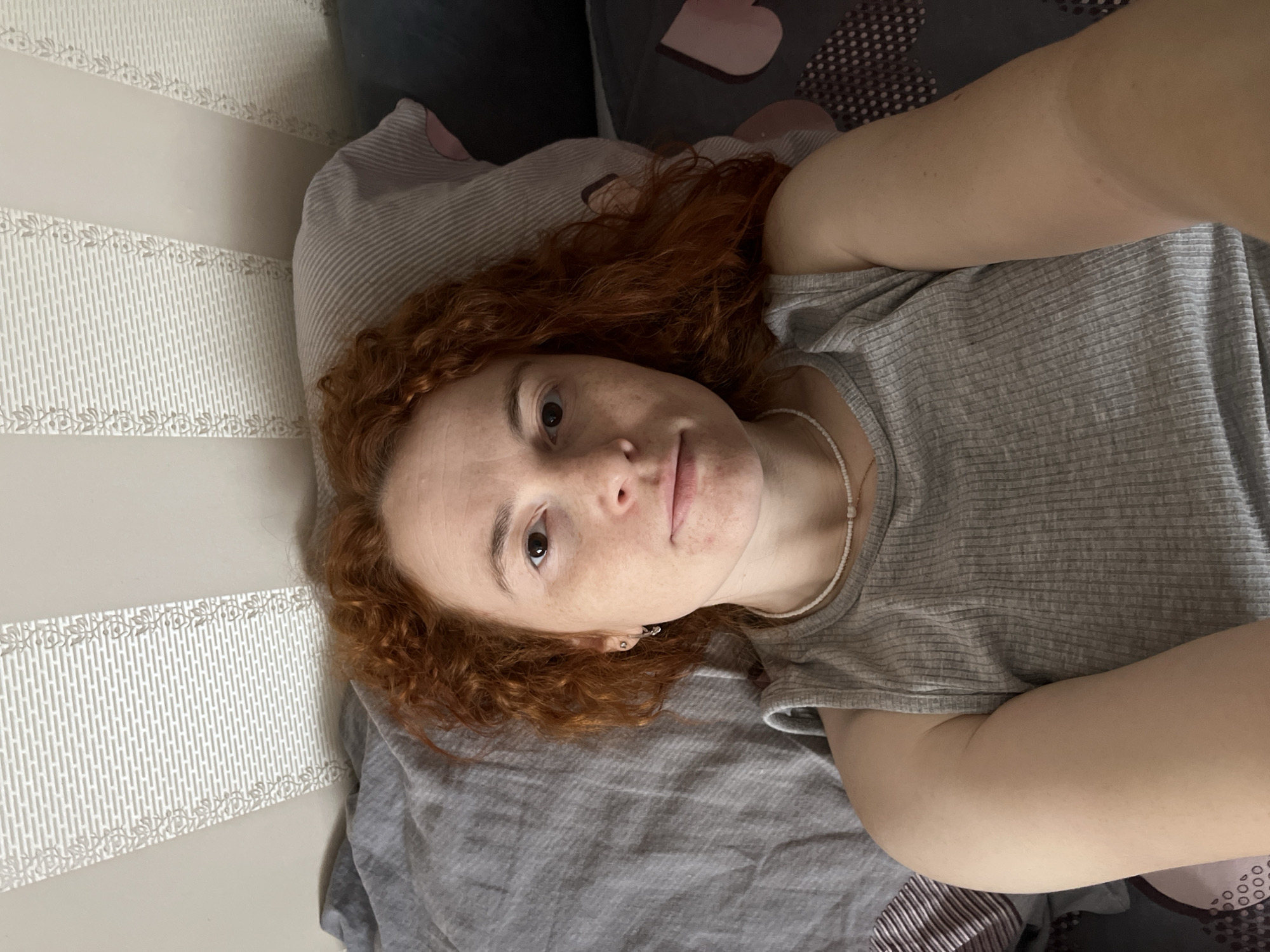 A selfie of a fair-skinned person with red curly hair, lying on a bed. The person is wearing a gray tank top and a beaded necklace. The background includes a gray pillow, striped bedding, and a patterned wall. The location is Chaikovsky, Russia.
