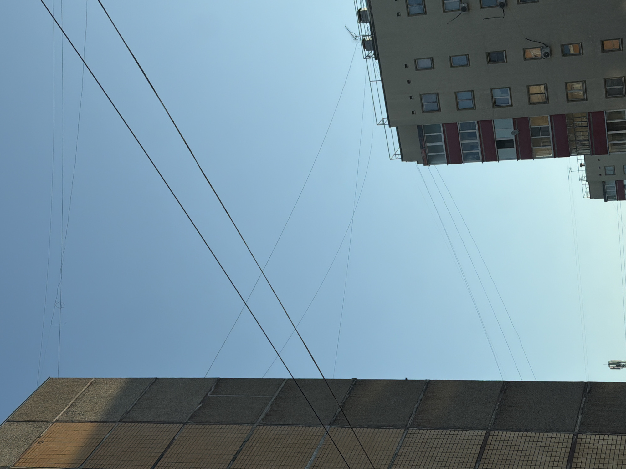 This image depicts an urban scene in Chaikovsky, Russia, looking upwards at buildings and utility lines against a clear, light blue sky. In the foreground, the top portion of a low-rise building with a patterned facade of beige and grey squares is visible. Above this building, crossing diagonally, are multiple black power lines that intersect. In the top right of the image, a taller multi-story building is visible, showing multiple rows of windows. Some windows have red framing or are open. There are also external air conditioning units attached to the building's facade. An antenna or satellite dish is also discernible on the building. The angle of the photograph suggests it was taken from street level, with the camera pointed towards the sky.