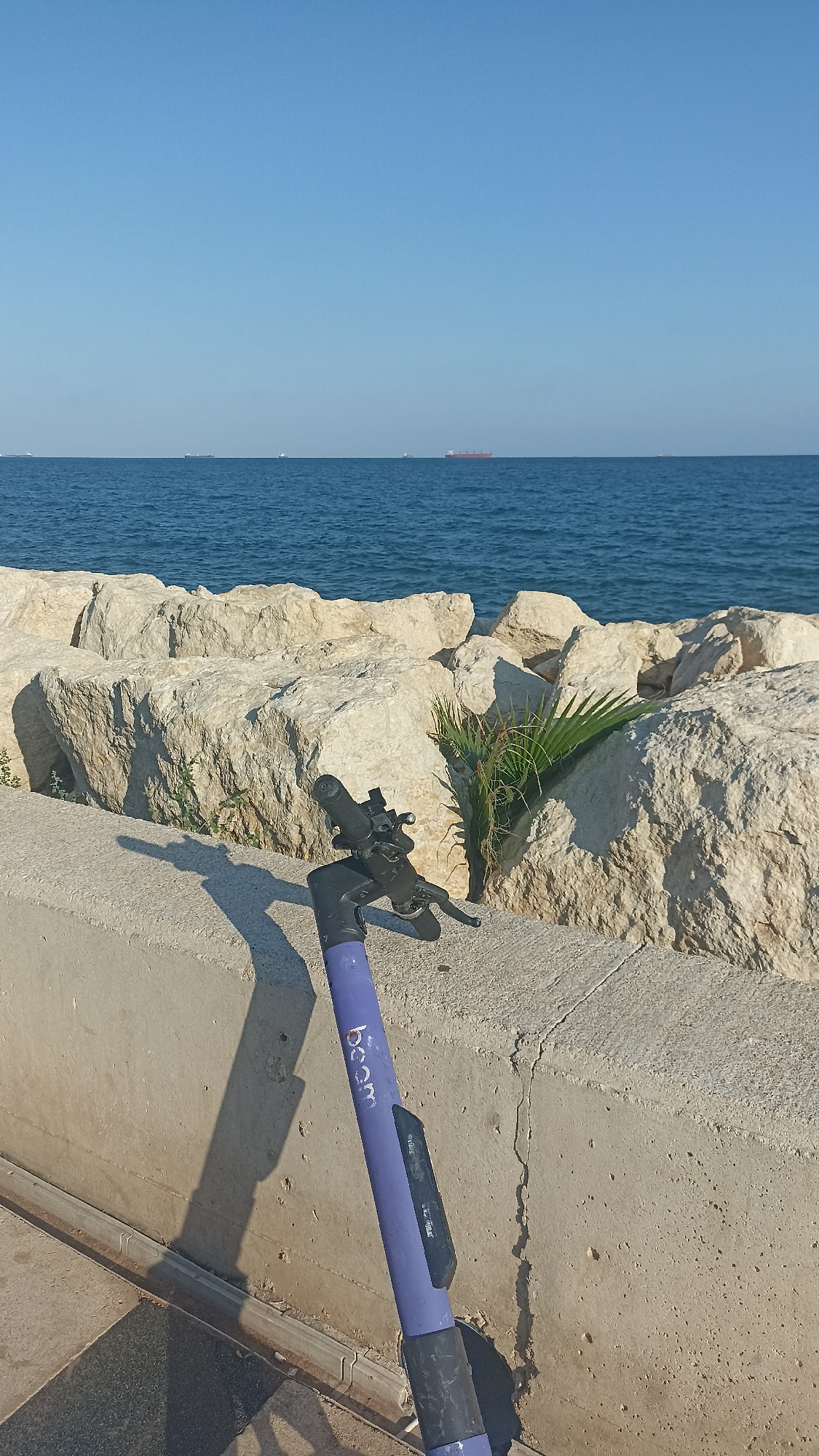 A purple electric scooter is parked on a concrete walkway overlooking the sea. The scooter is angled towards the viewer, with its handlebars and front wheel visible. The word "beam" is visible in white lettering on the scooter's frame. Behind the scooter, a wall of large, light-colored rocks lines the edge of the walkway. Beyond the rocks is the calm, blue sea, with several cargo ships visible on the horizon. The sky is a clear, bright blue, suggesting a sunny day. The sun casts a long shadow of the scooter onto the walkway. A small palm frond is visible growing amongst the rocks. The scene appears to be a coastal area in Yenişehir, Turkey, likely during the daytime.