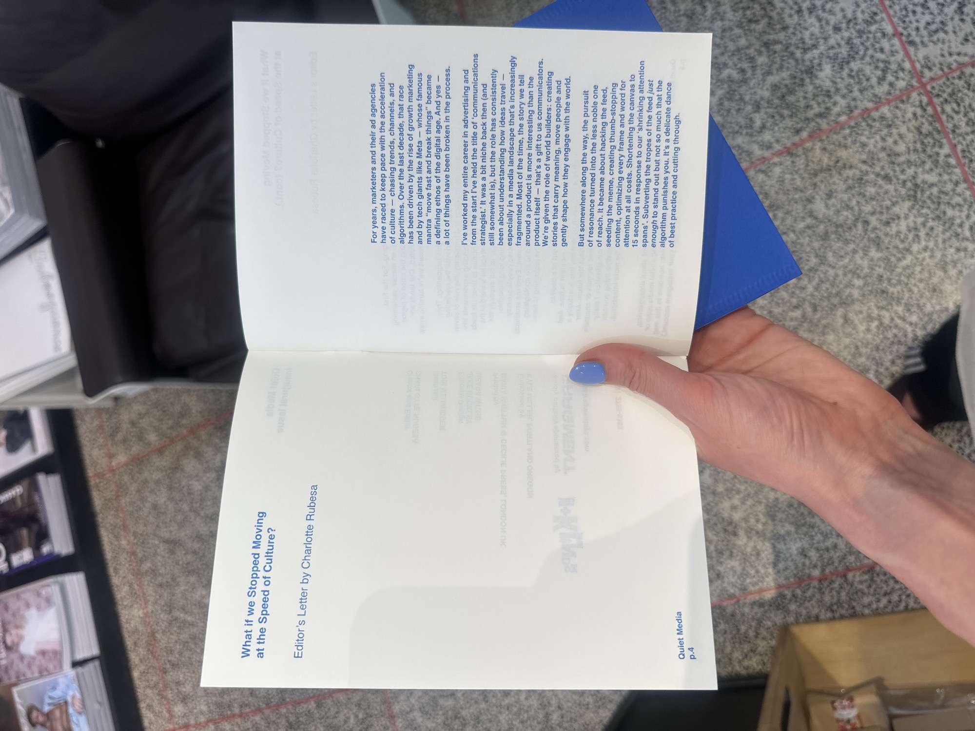 The image shows a person holding open a booklet or small magazine. The left-hand page has the title "What if we Stopped Moving at the Speed of Culture?" and "Editor's Letter by Charlotte Rubesa," with "Quiet Media p.4" at the bottom. The right-hand page contains text in columns, discussing marketing, advertising, and the impact of digital culture on communication. The person's hand is visible, with light blue nail polish. The background includes other copies of the booklet, and the surface has a mottled, patterned texture. The lighting is bright and appears to be artificial. It is unlikely that the location is in London, United Kingdom as nothing is in the picture to support that.