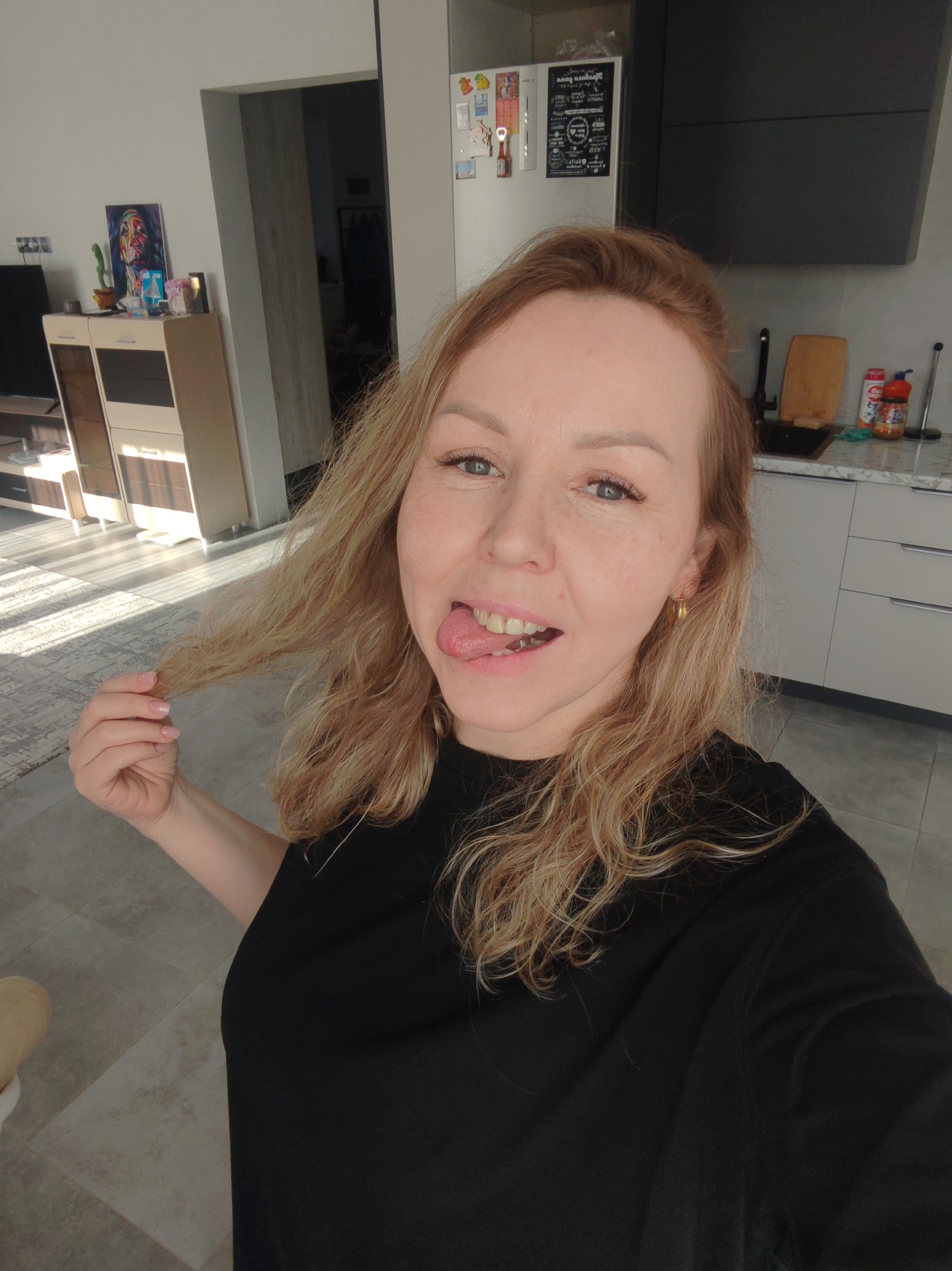 A woman with blonde hair, wearing a black shirt and gold earrings, is posing for a selfie in an indoor environment. She has her tongue out and is holding a strand of her hair in her left hand. The background includes a kitchen area with white cabinets, a countertop with items like a cutting board and food containers, and a black range hood. Adjacent to the kitchen is a living area with a light-colored wooden cabinet, a TV, and a gray patterned rug. The image was taken in a location identified as the city of Chaikovsky, Russia.