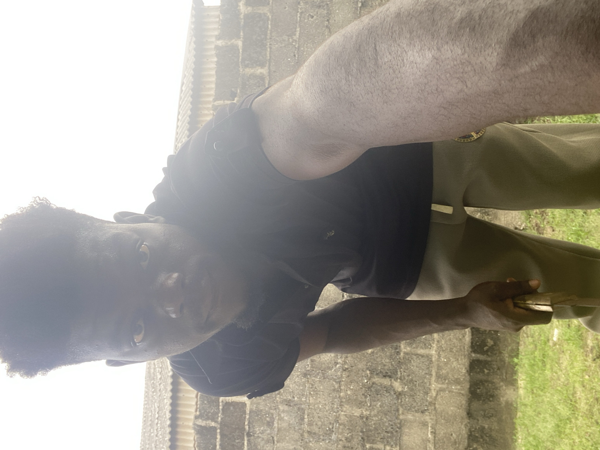 A man with dark hair and skin is photographed from a low angle, against a backdrop of a concrete block wall on one side and green grass on the other. He is wearing a black t-shirt and olive green pants with a decorative emblem on the upper thigh. In his hand, he holds what appears to be a flat, wooden object. The location is likely outdoors in an urban setting in the city of Unknown, Unknown.