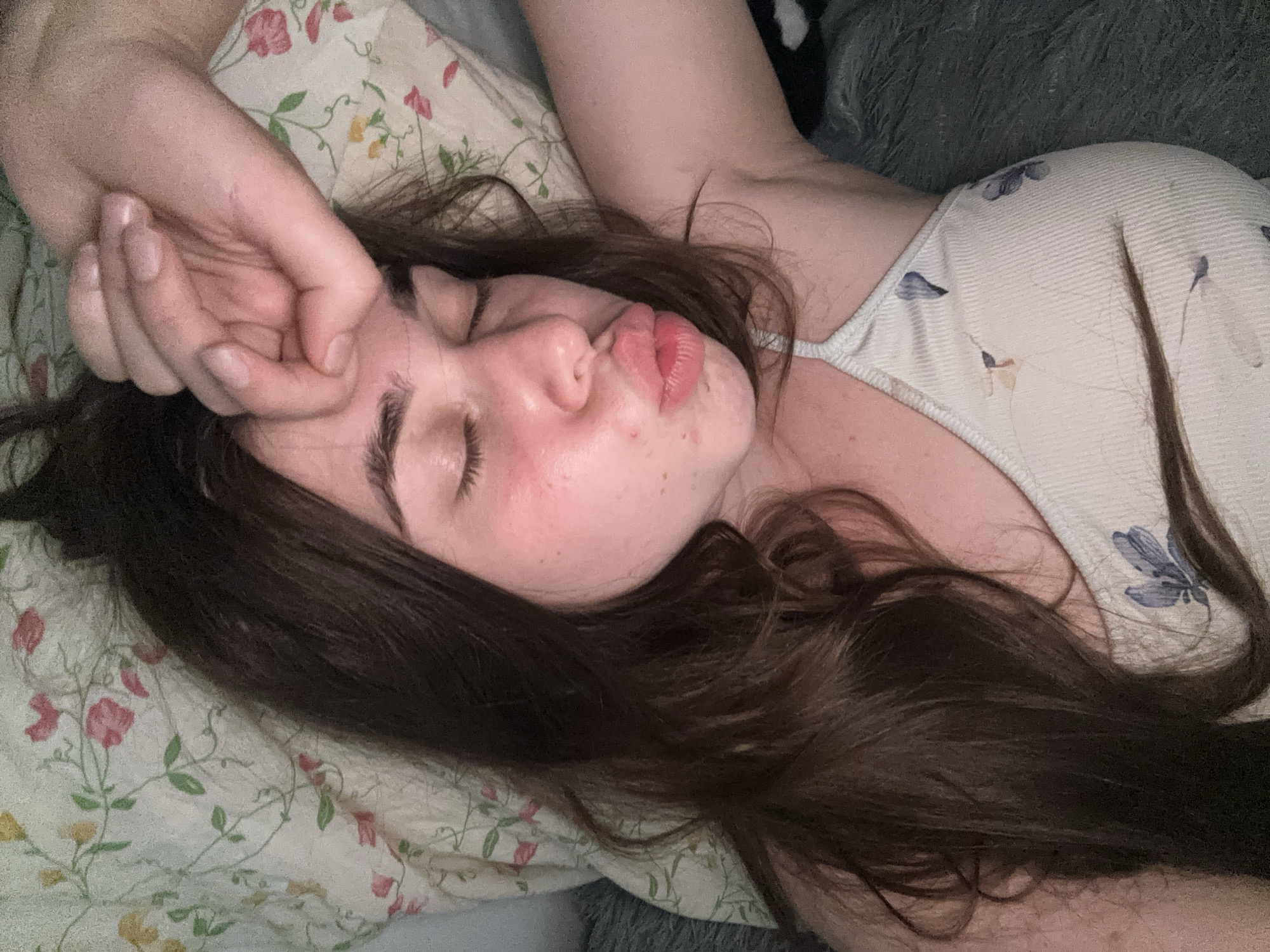 The image shows a young woman with her eyes closed, lying on her side on a floral-patterned pillow. Her right hand is raised to her forehead. Her lips are pursed, as if she is about to kiss or is pouting. She is wearing a light-colored camisole with a floral print. The background appears to be a bed with a dark, furry throw blanket. The overall impression is of someone resting or sleeping. There is no indication of the location being in the city of Мещёры, Russia.