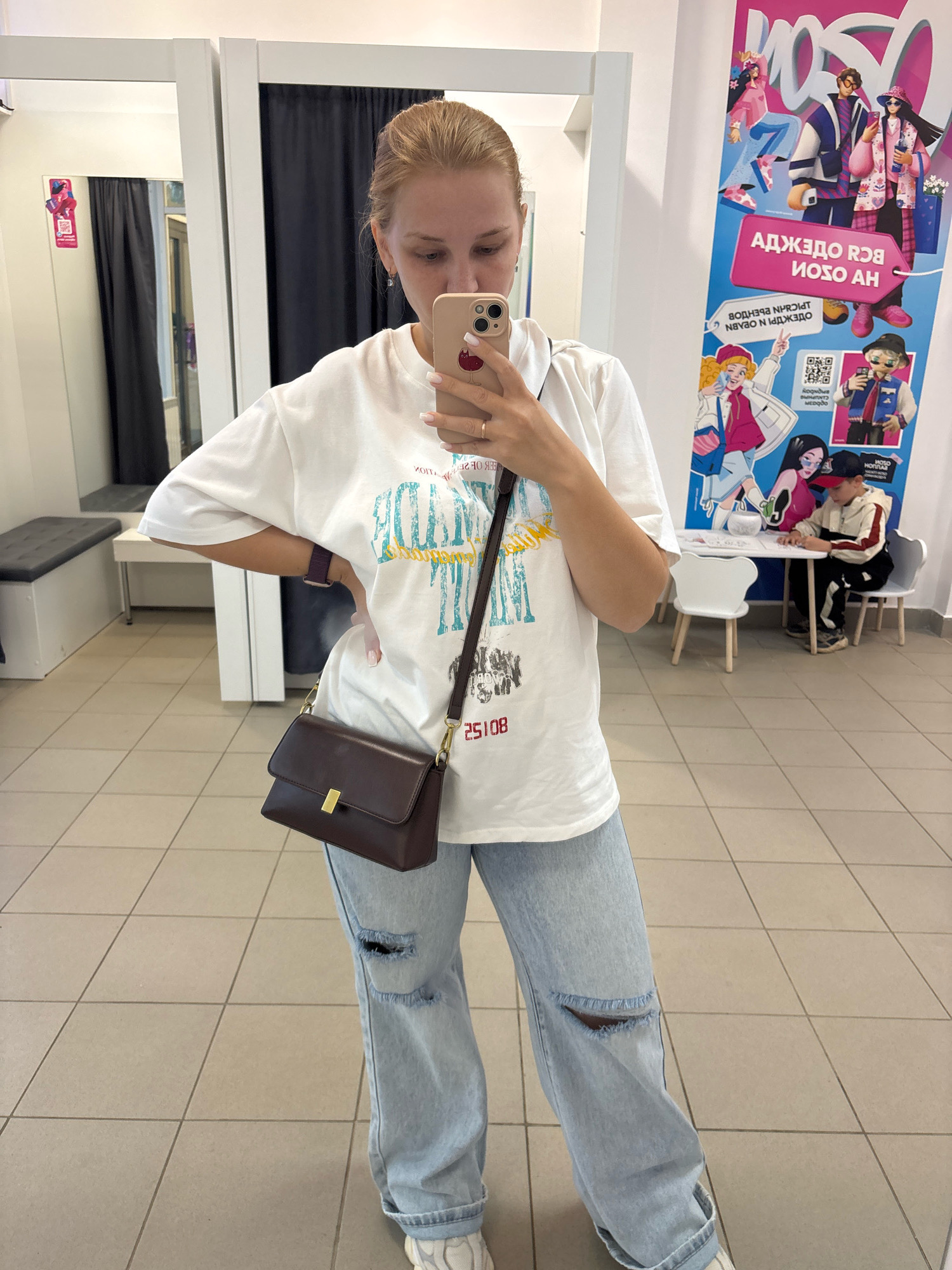 A young Caucasian woman with blonde hair pulled back stands in what appears to be a fitting room or retail space, taking a mirror selfie. She is wearing a white oversized t-shirt with a graphic print, light-wash distressed denim jeans, and white sneakers. She carries a dark brown crossbody bag with a gold clasp. Her left hand holds a smartphone, and her right arm is bent at the elbow with her hand resting on her hip. In the background, a large colorful poster with animated characters is visible, along with fitting room curtains and tiled flooring. A young boy is seated at a small table in the background. The environment is consistent with a retail store, possibly in Kuznechikha, Russia, judging by the Cyrillic text on the poster.