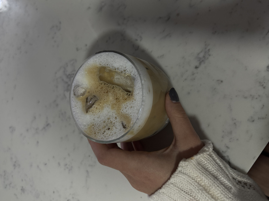 An overhead view depicts a fair-skinned human hand, partially clad in a white ribbed knit sweater sleeve, holding a clear glass of iced coffee. The beverage consists of a light brown liquid, topped with a thick layer of white foam and darker brown liquid drizzles, containing several irregular ice cubes. The thumb, displaying dark nail polish, is visible gripping the glass. The scene is set indoors on a light-colored countertop featuring subtle gray and black mottled patterns, in the city of Unknown, Unknown.