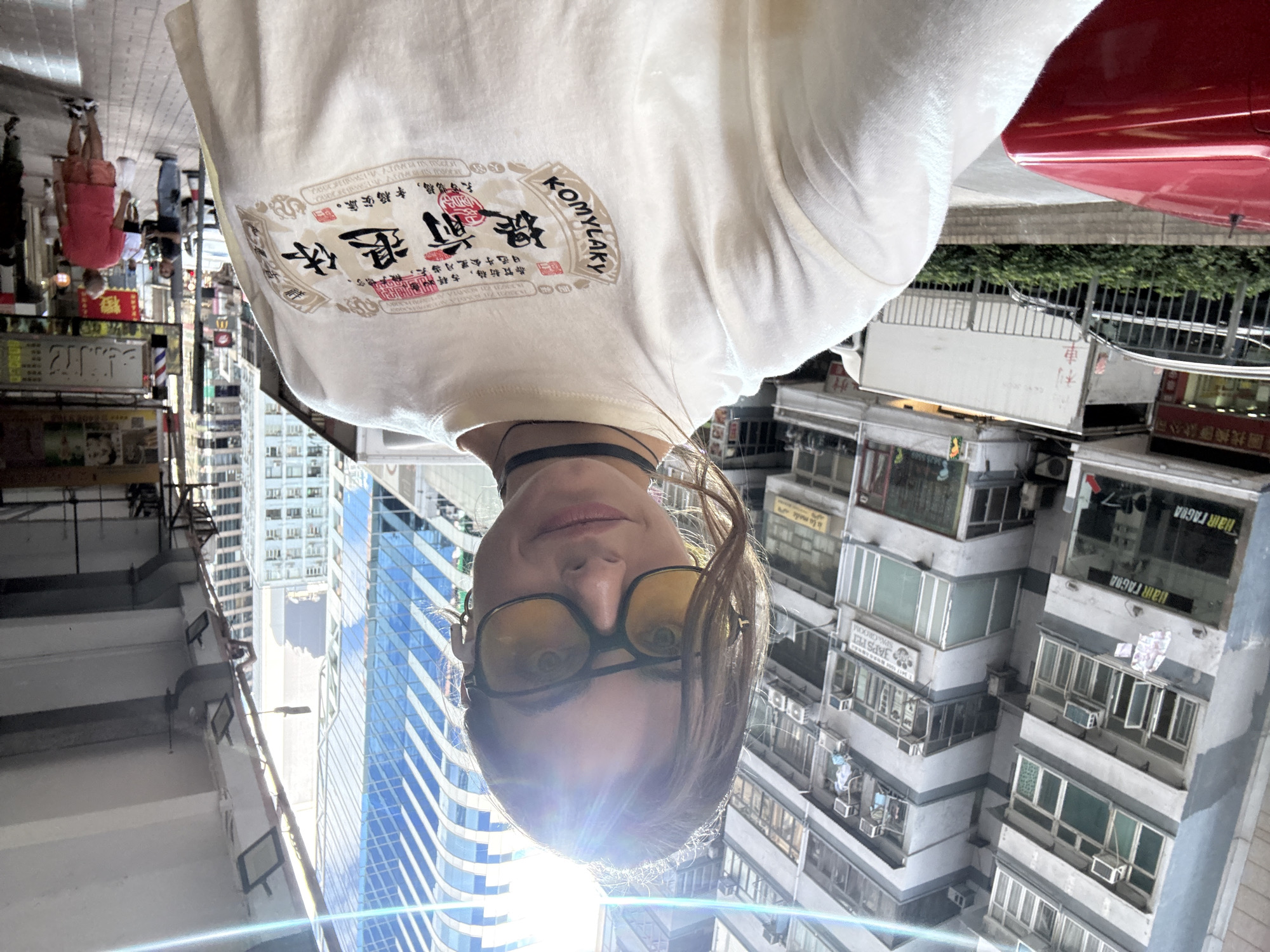 The image depicts a person wearing a white T-shirt with a graphic design and yellow-tinted sunglasses, seemingly taking a selfie with a backdrop of urban scenery.
The background shows a typical street in Hong Kong, China, with densely packed buildings, a mix of old and new architecture, including a modern high-rise skyscraper. A red vehicle is visible on the right side of the image. There's also a street scene with people and shops, albeit out of focus. The environment suggests a bustling, urban area.