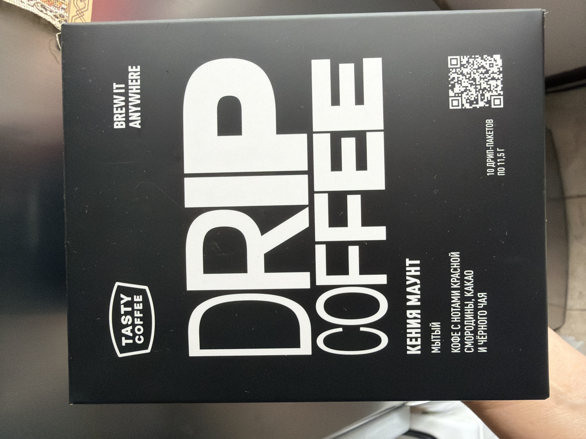 The image shows the front of a black box of "TASTY COFFEE DRIP COFFEE." The box features large white lettering. The top left corner has the "TASTY COFFEE" logo. The top right corner has the text "BREW IT ANYWHERE." Below the "DRIP COFFEE" text, there is Russian text that translates to "KENYA MOUNT," "Washed," and "COFFEE WITH NOTES OF RED CURRANT, COCOA AND BLACK TEA." The bottom right corner has a QR code and text indicating "10 DRIP-PACKETS OF 11.5 G." The box is lying on a surface, partially obscured by a person's hand. The background appears to be a dark, possibly indoor environment. No specific details about the city of Khimki, Russia, are visible in the image.